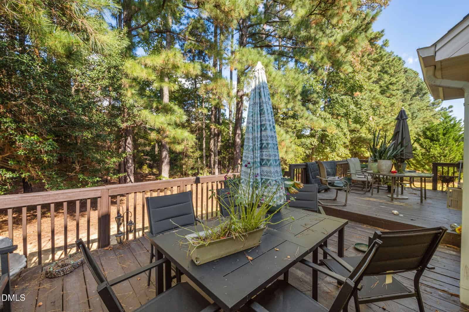 A wooden deck with two patio tables, black chairs, potted plants, and two closed umbrellas. Trees and greenery surround the deck, and sunlight filters through the branches.