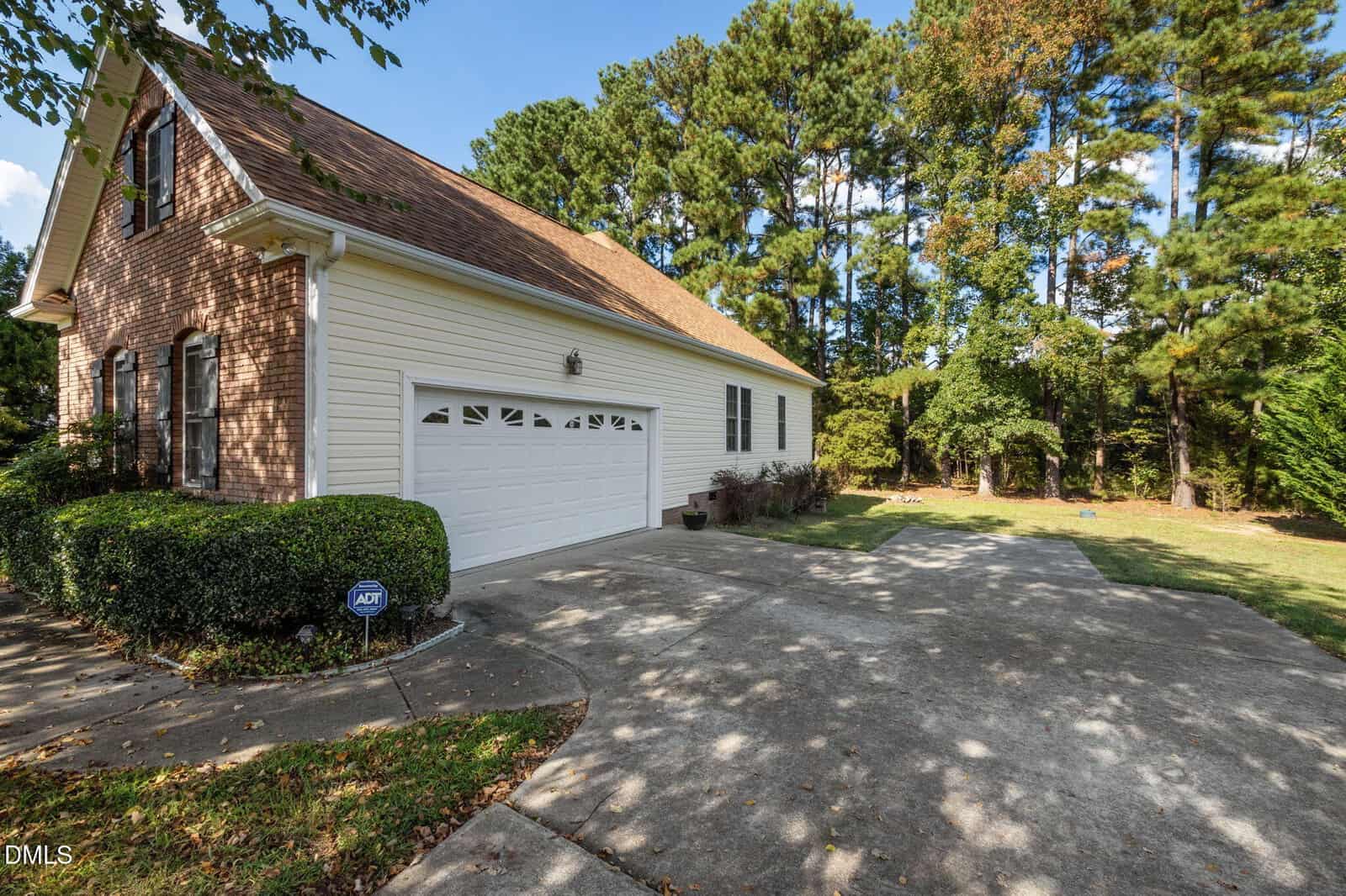 A two-car garage attached to a house with brick and siding exterior, surrounded by trees and shrubs, with a concrete driveway and security sign in front.