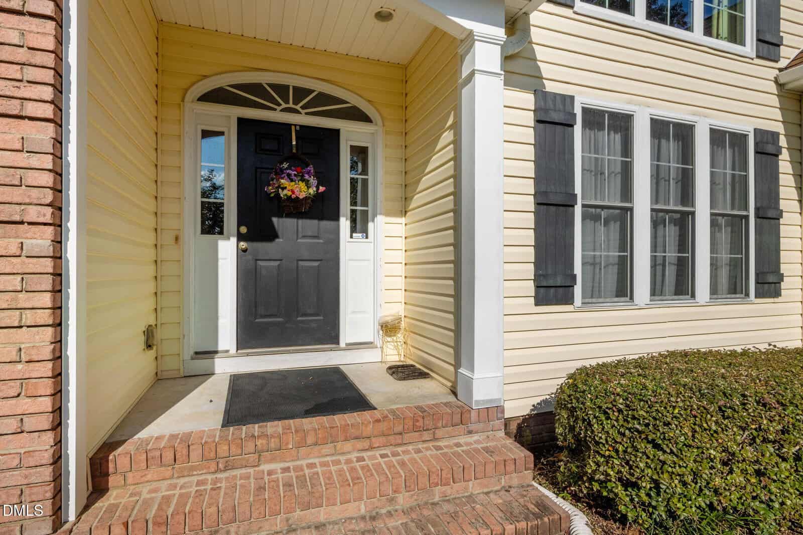 A black front door with a floral wreath opens to a small porch with brick steps, white columns, and yellow siding. Three tall windows with black shutters are on the right, and bushes line the walkway.