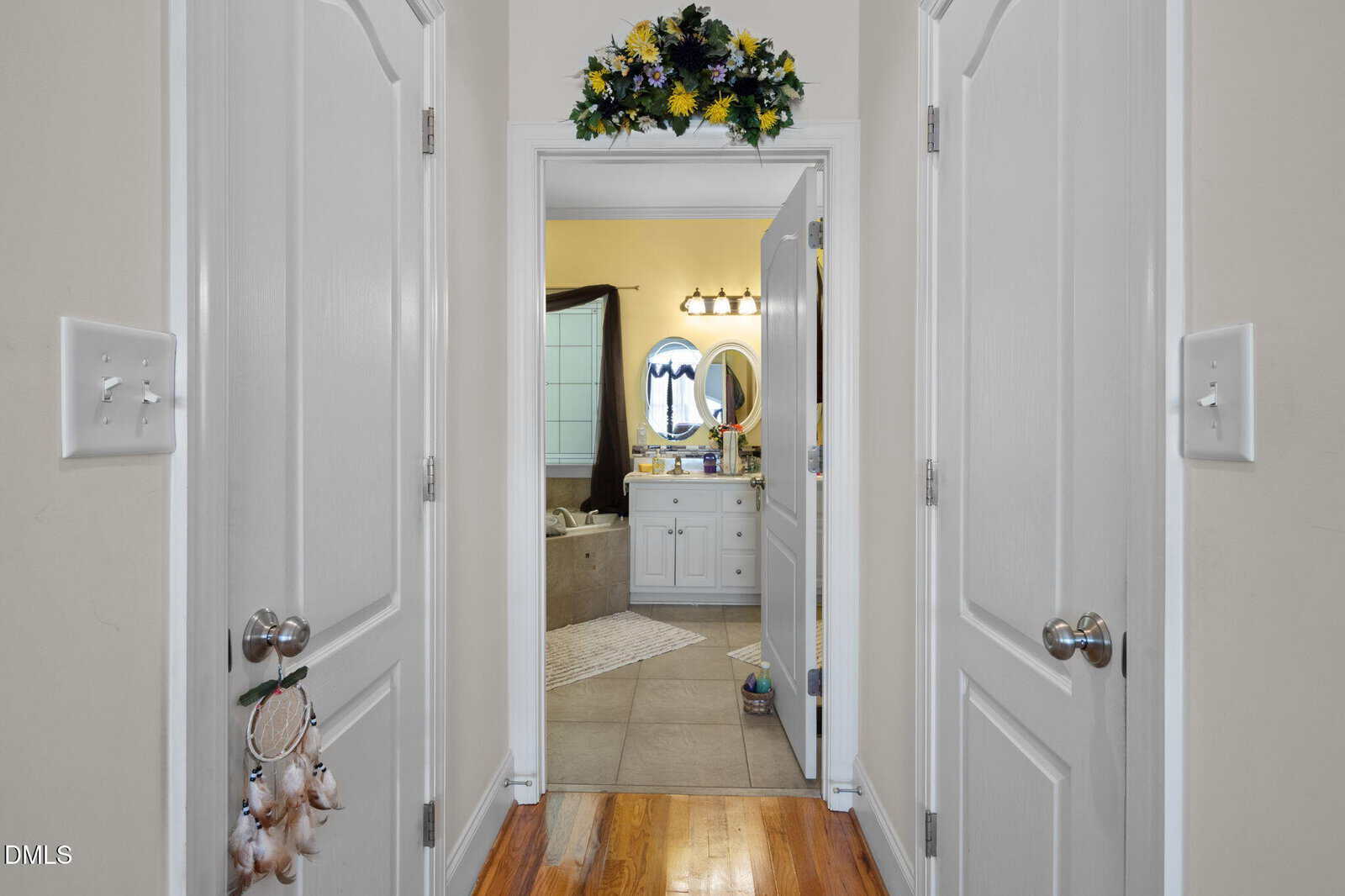 View through two white double doors into a bathroom with a white vanity, round mirror, and flower arrangement above the doorway; wooden floor in the hallway, tile floor in the bathroom.