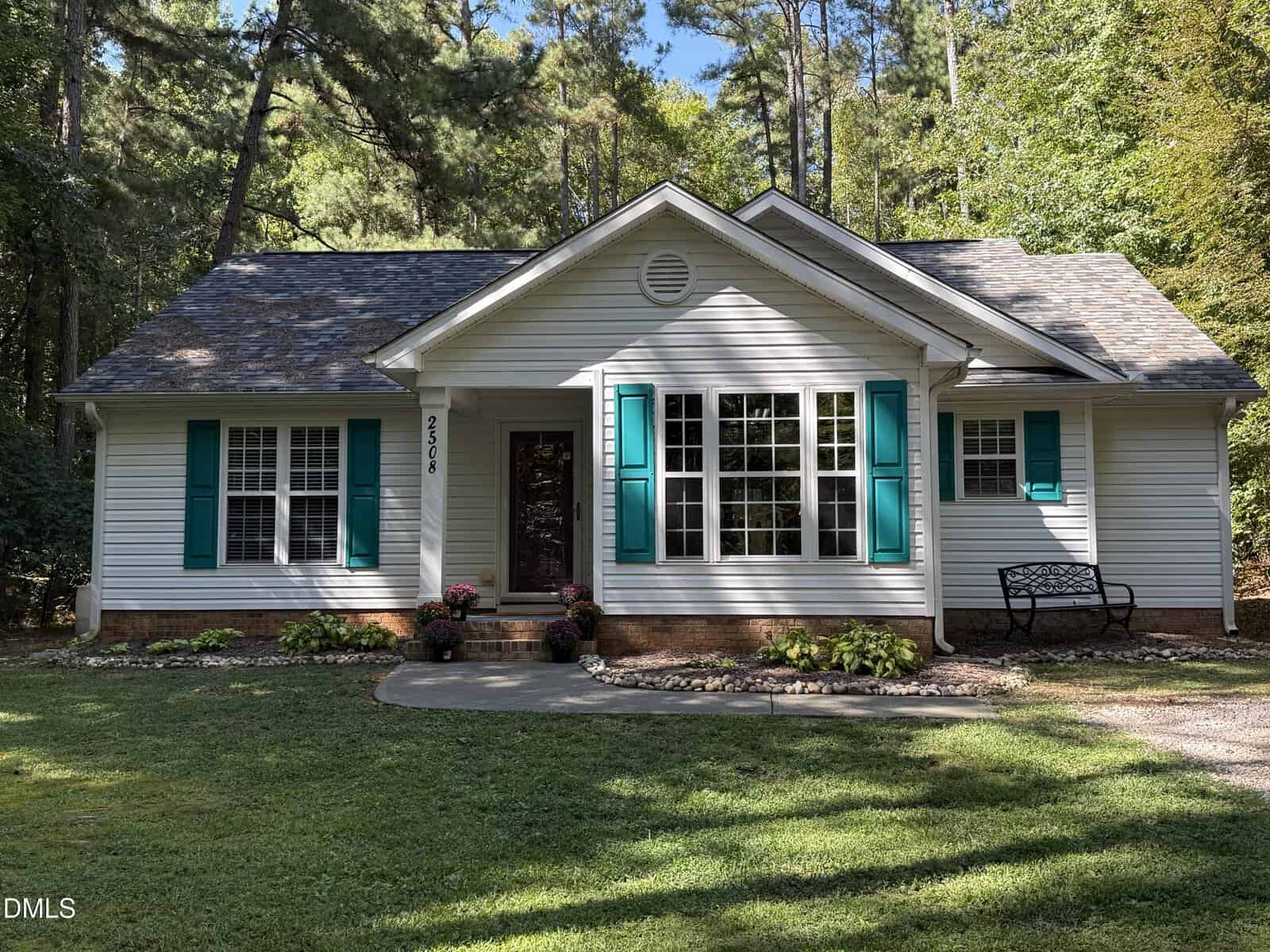 Charming single-story white house at 2508 Patton Lane in Franklinton, featuring teal shutters, a central front door, and a small stoop. A black bench sits to the right amid trees and grass, with sunlight filtering through the leaves.