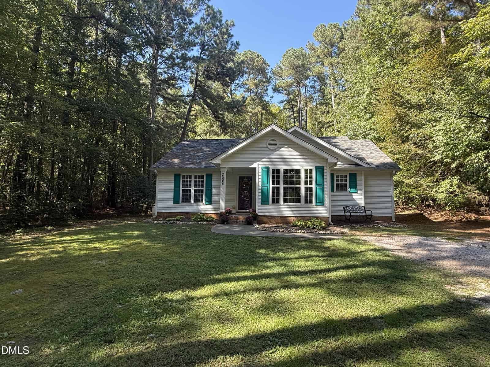 A small white house with teal shutters and a shingled roof sits in a grassy yard surrounded by tall trees. A curved path leads to the front door, with a bench and flowerbed near the entrance.