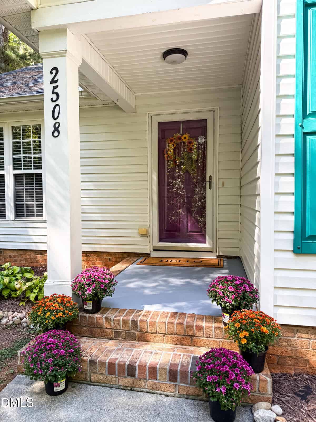 Front porch of a house with the number 2508 on a post. The door is painted purple and decorated with a sunflower wreath. Potted purple, orange, and pink flowers are arranged on the brick steps and landing.