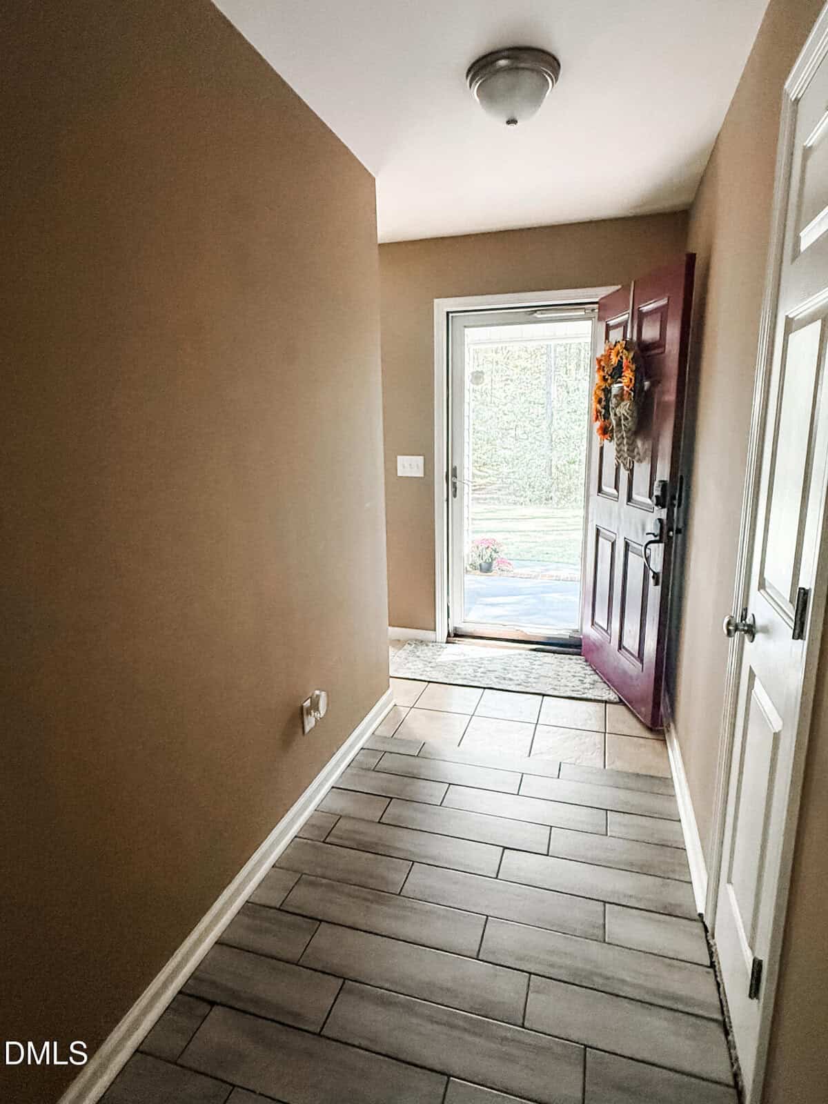 A hallway with tan walls and light-colored tile flooring leads to an open front door. The door is red and has a fall-themed wreath hanging on it. Daylight streams in from outside.
