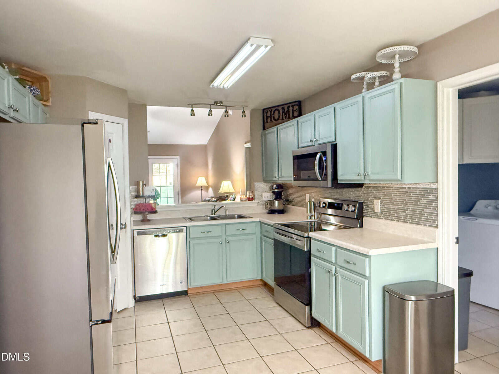 A kitchen with light blue cabinets, stainless steel appliances, beige countertops, tiled floor, and a white ceiling. There is a dishwasher, stove, microwave, refrigerator, and a trash can visible.