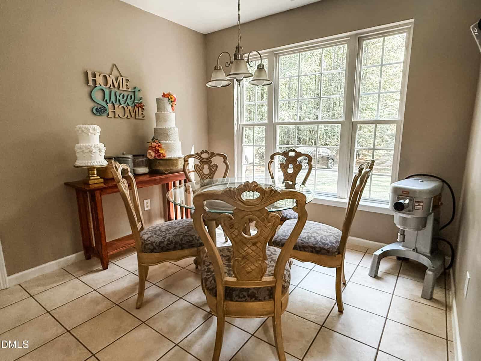 Bright dining area with a round glass table, four ornate chairs, cakes on stands, “Home Sweet Home” wall decor, large windows, and tiled floor. A stand mixer sits by the window.