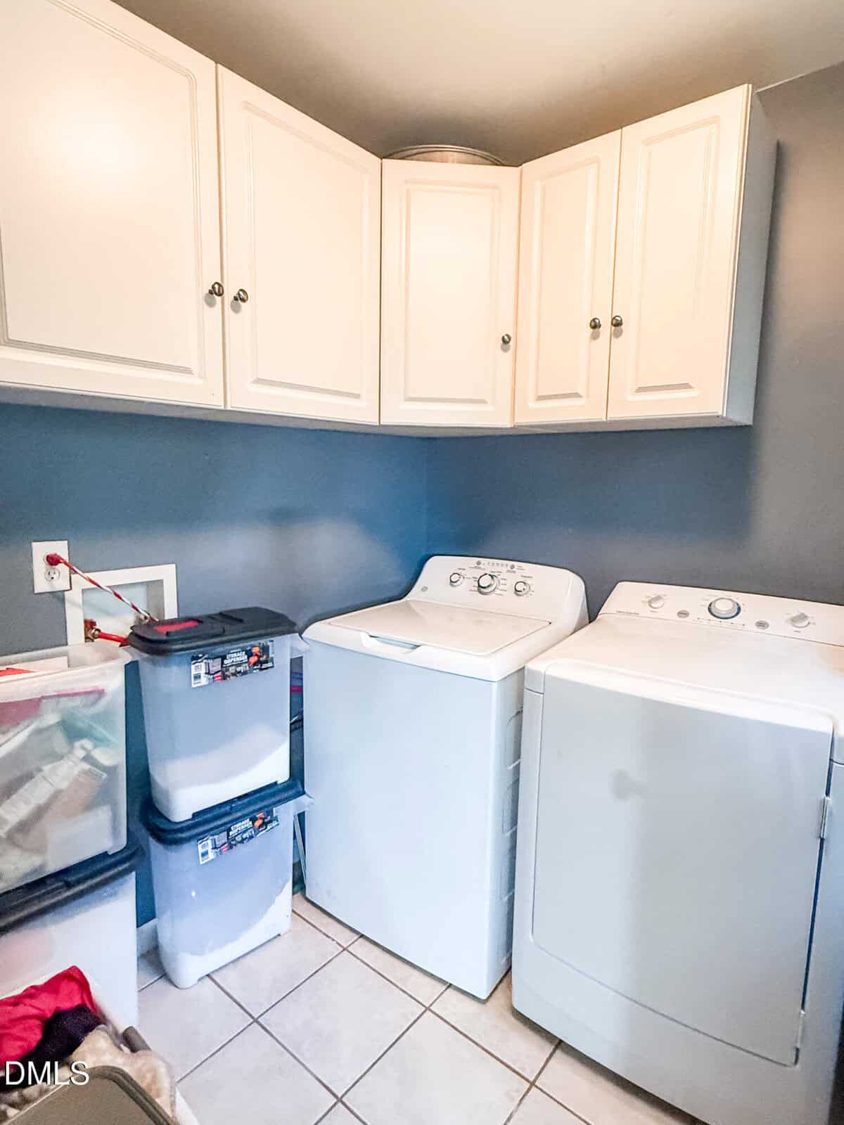 A laundry room with a white washing machine and dryer side by side, white cabinets above, two stacking plastic storage bins, a basket with laundry items, and gray walls.