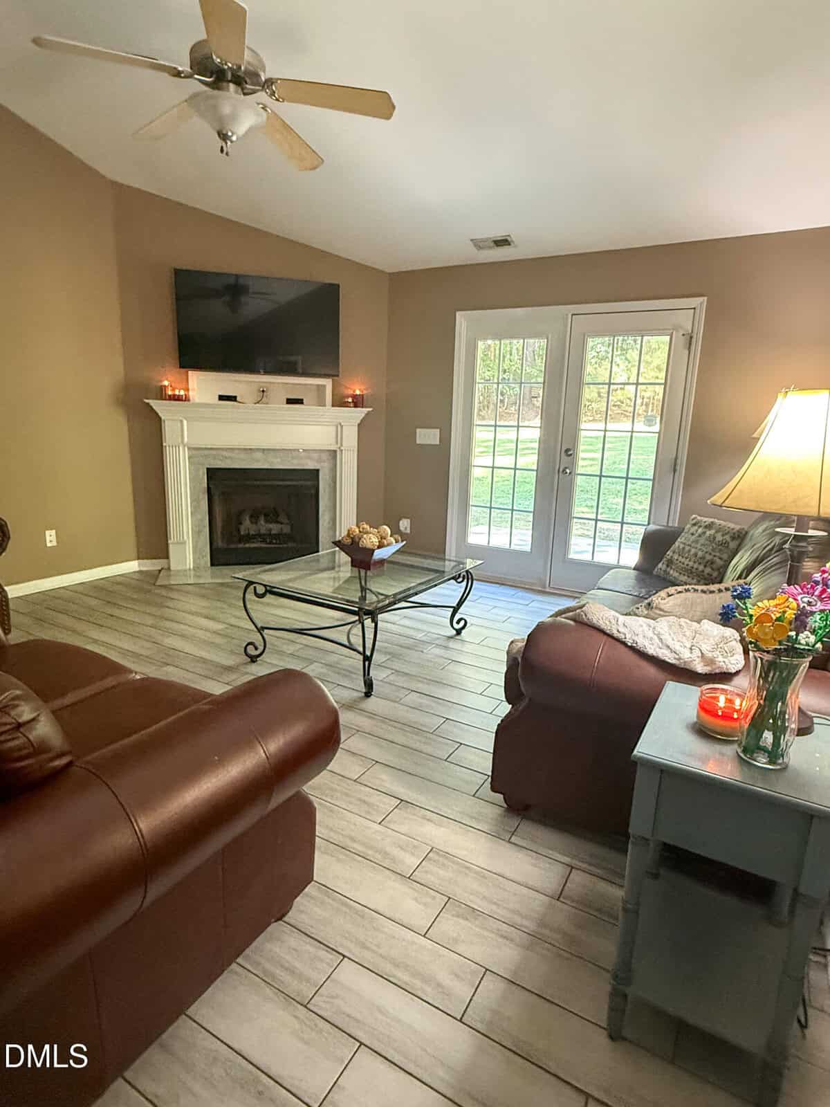 A living room with two brown leather sofas, a glass coffee table, a fireplace with a TV above it, a side table with a lamp and flowers, and glass double doors leading outside. The floor is light wood-style tile.