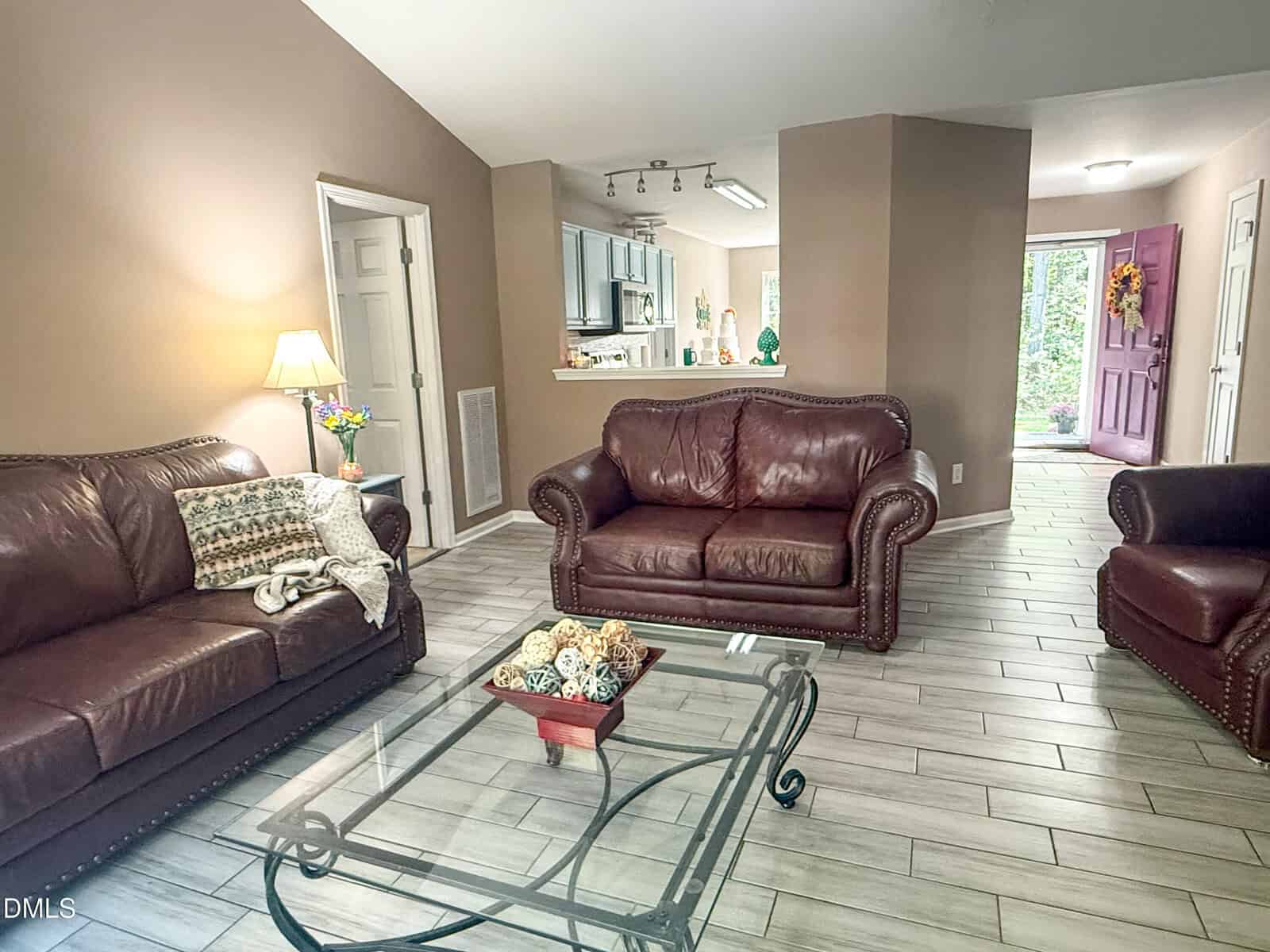 A cozy living room with brown leather sofas, a glass coffee table, and a lamp. The open layout leads to a kitchen and a hallway with a purple front door decorated with a wreath.