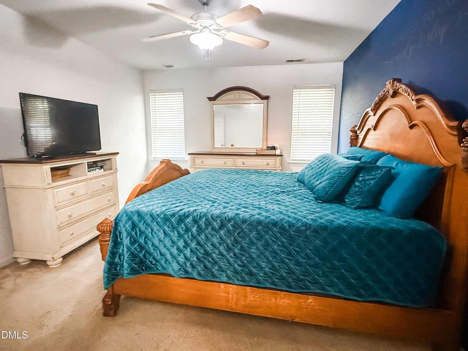 Bedroom with a wooden bed frame, blue quilt and pillows, two white dressers, a flat-screen TV, a mirror, and two windows. A ceiling fan hangs above the bed. The accent wall behind the bed is blue.