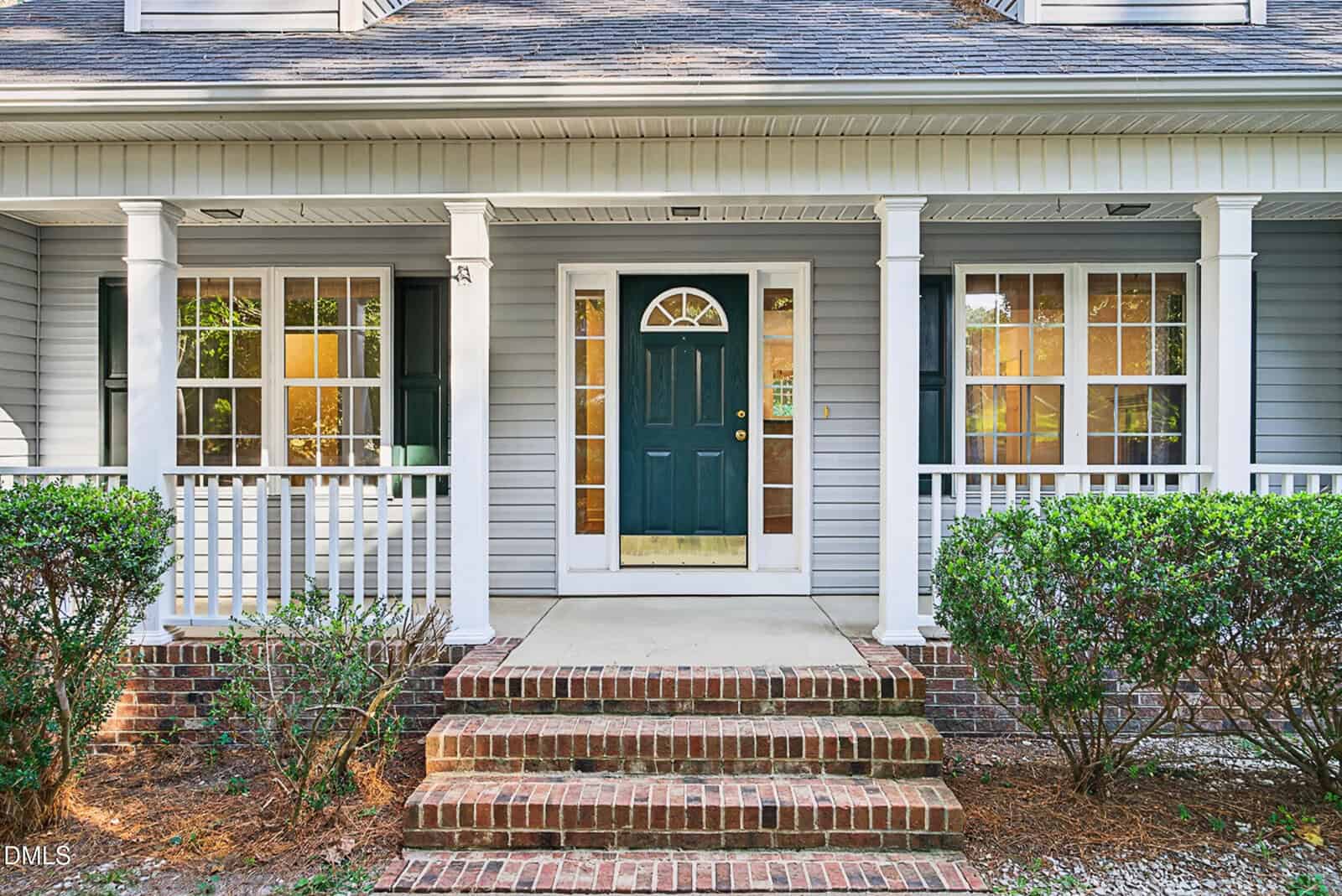 Front view of a house with a green door, sidelights, two windows, white columns, a small porch, brick steps, and shrubs on either side of the entrance.