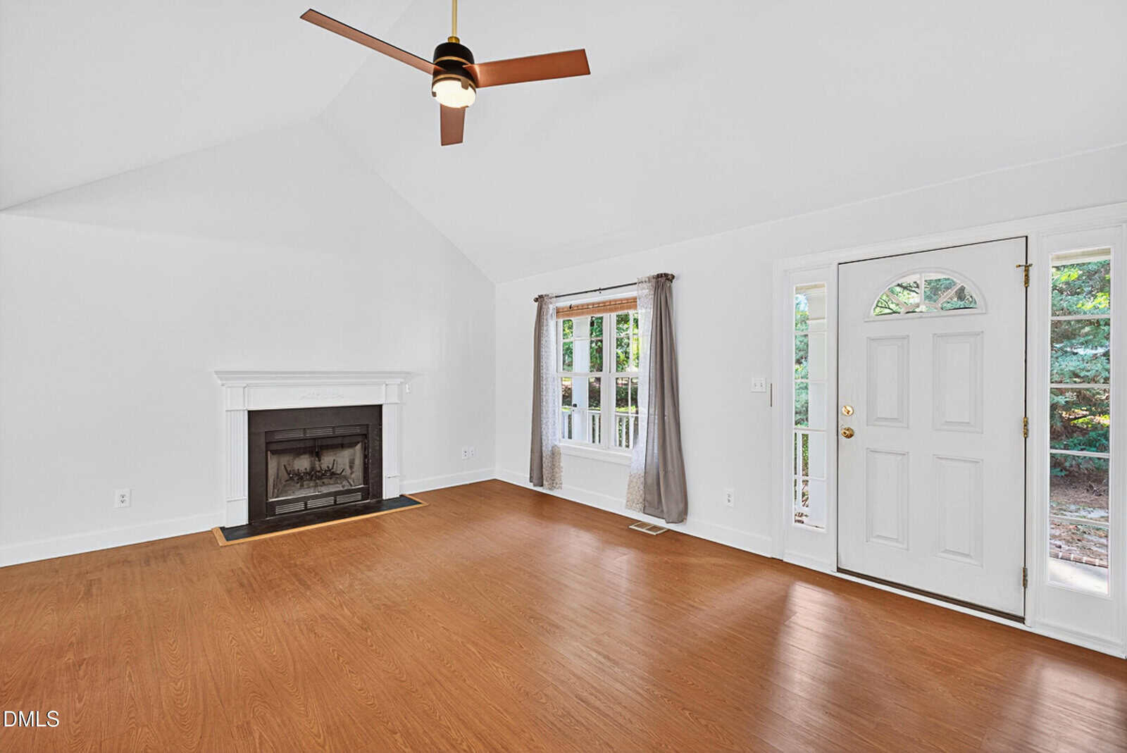 A living room with white walls, wood flooring, a ceiling fan, a fireplace with a white mantle, a window with gray curtains, and a white front door with glass panels. Trees are visible outside the window and door.