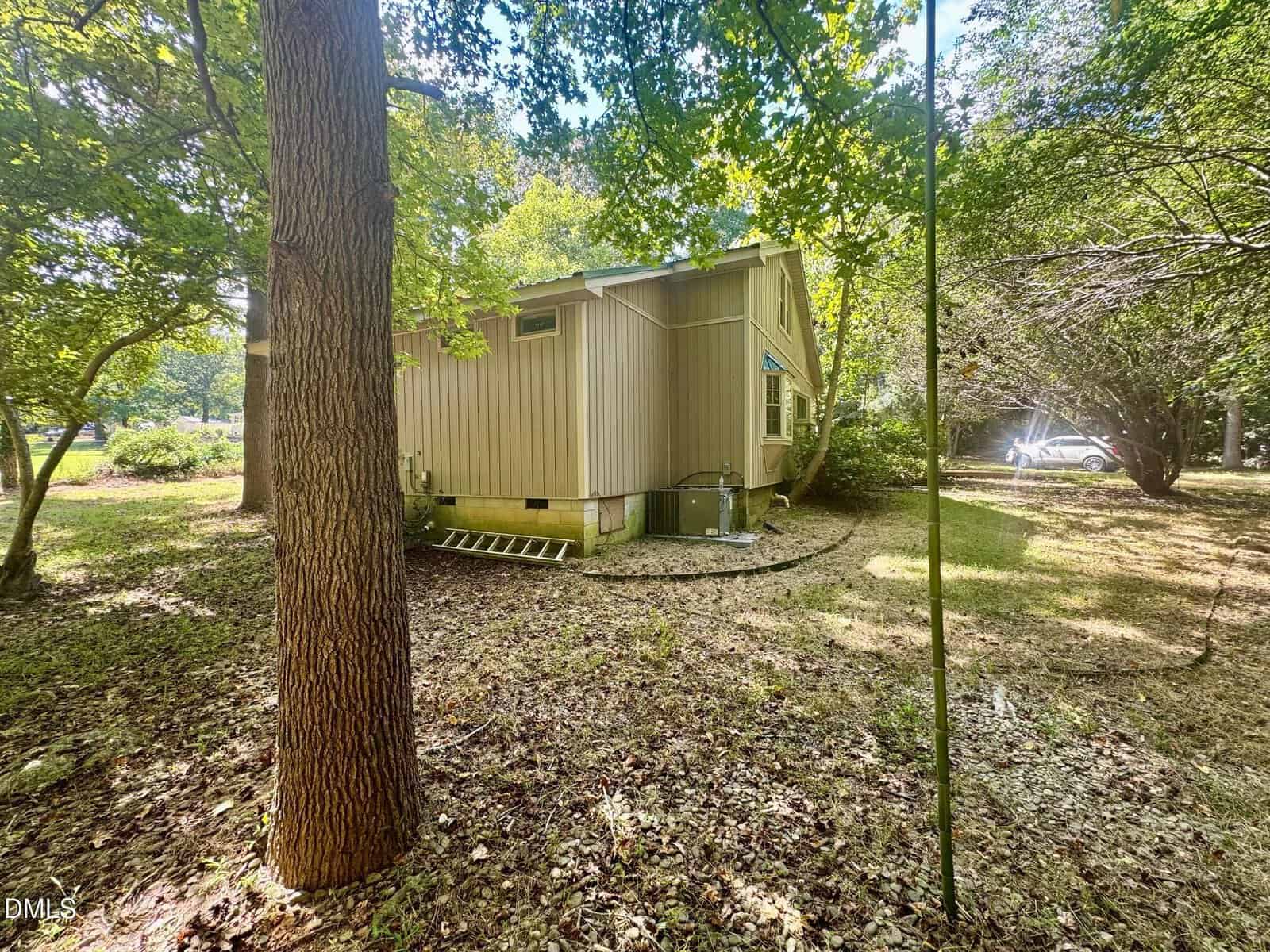 A beige house with vertical siding sits among trees. There is a metal ladder on the ground beside the house, and a car is partially visible in the background near bushes. The yard is covered with dry leaves.