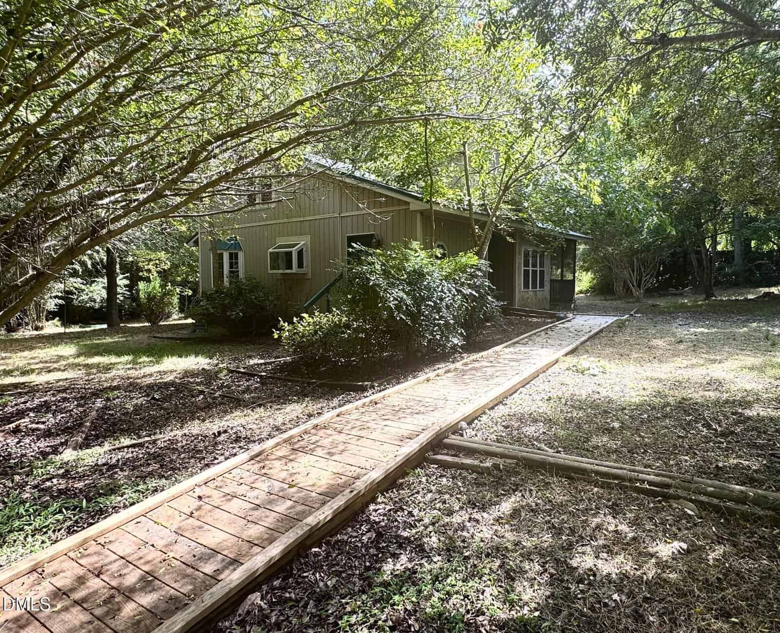 A wooden walkway leads to a small, beige house with green trim, partially shaded by trees and surrounded by bushes and grass. Sunlight filters through the leaves, casting dappled shadows on the ground.