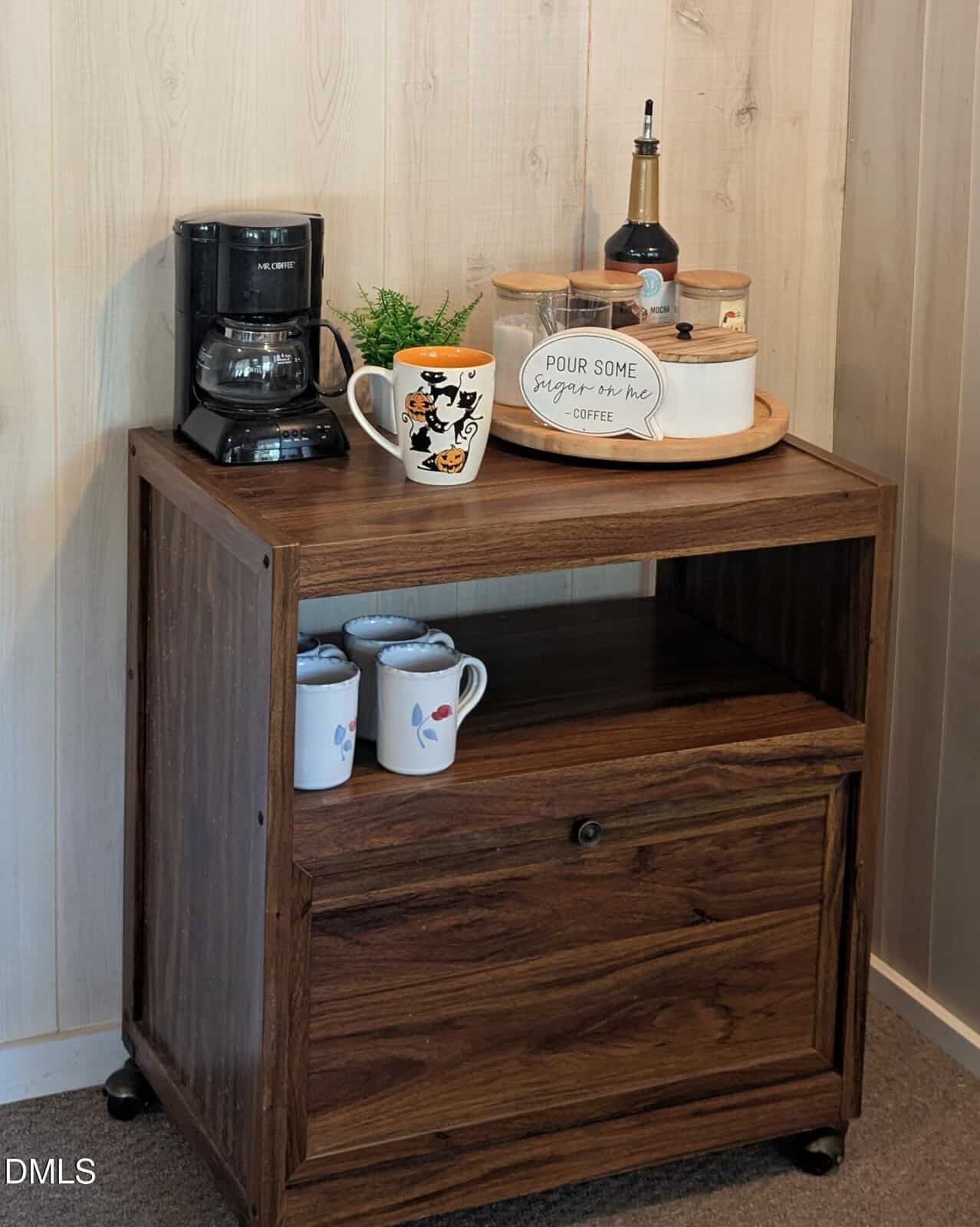 A small wooden cart with a coffee maker, mugs, creamer, and a coffee sign on top. The middle shelf holds additional mugs, and the bottom section is closed. The cart is set against a light wood-paneled wall.