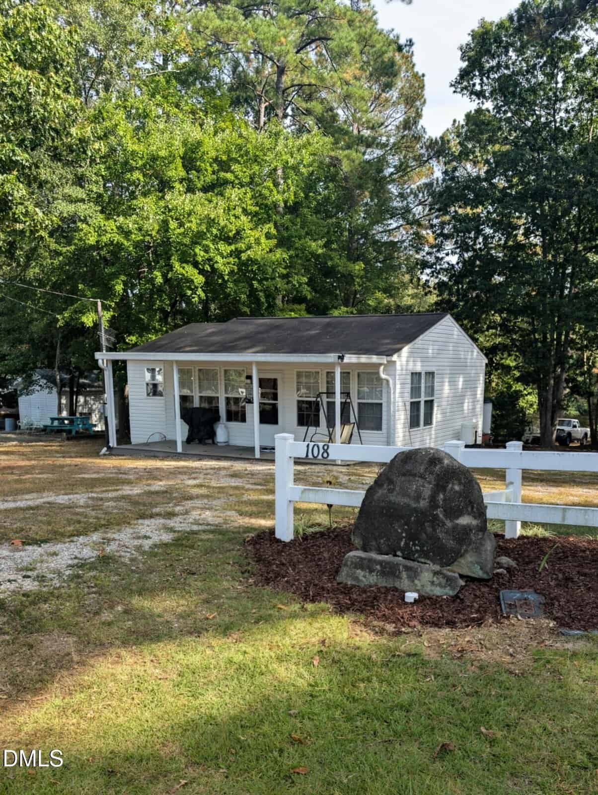 A small white house with a covered front porch sits behind a white fence at 108 Red Oak Drive, Louisburg. A large rock with mulch is in the foreground, and the yard is surrounded by tall green trees. The house number 108 is visible on the fence.