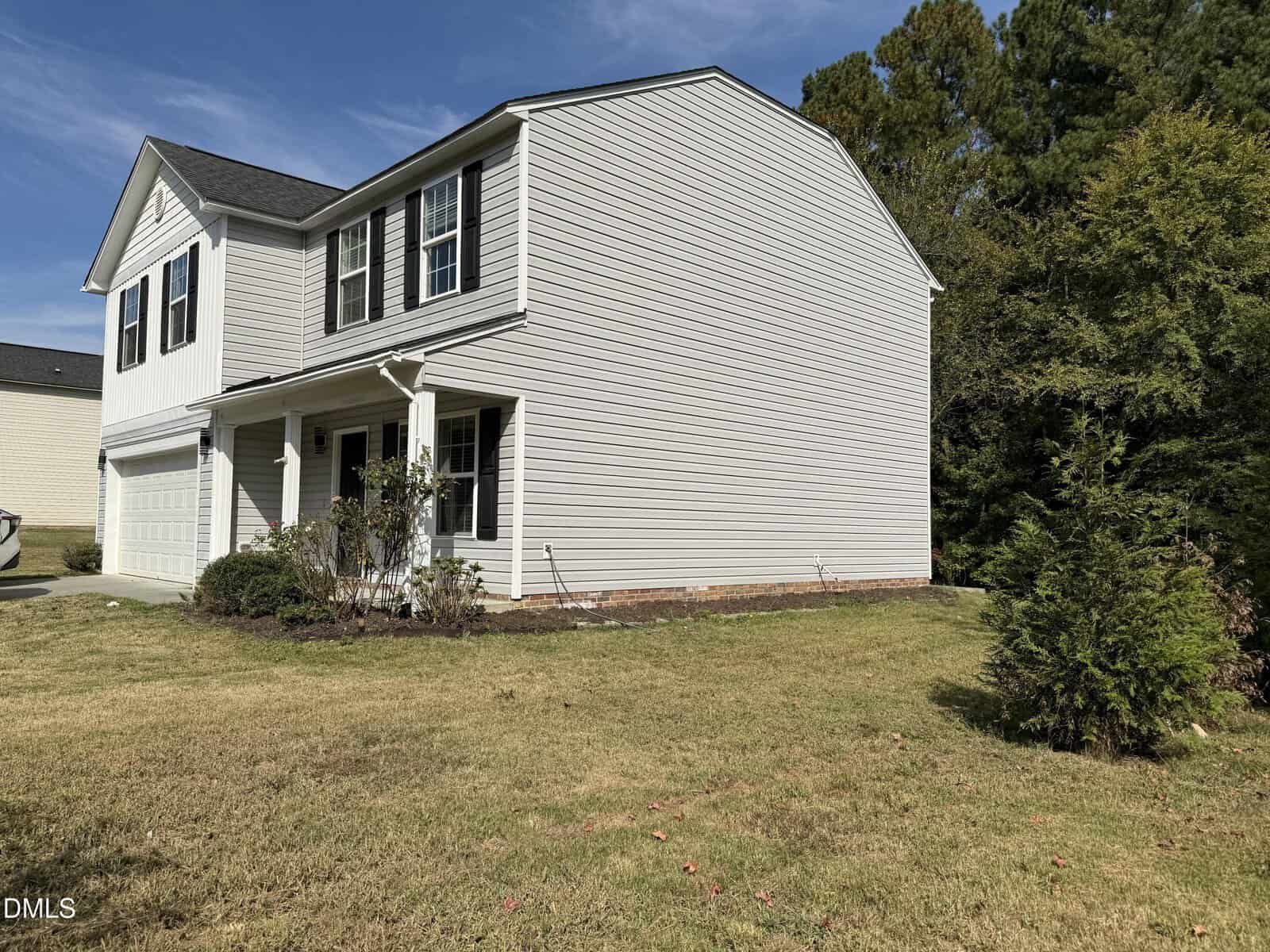 A two-story beige house with white trim, black shutters, and a covered front porch. The house has a two-car garage and sits on a grassy lawn with a few shrubs and trees around the property.