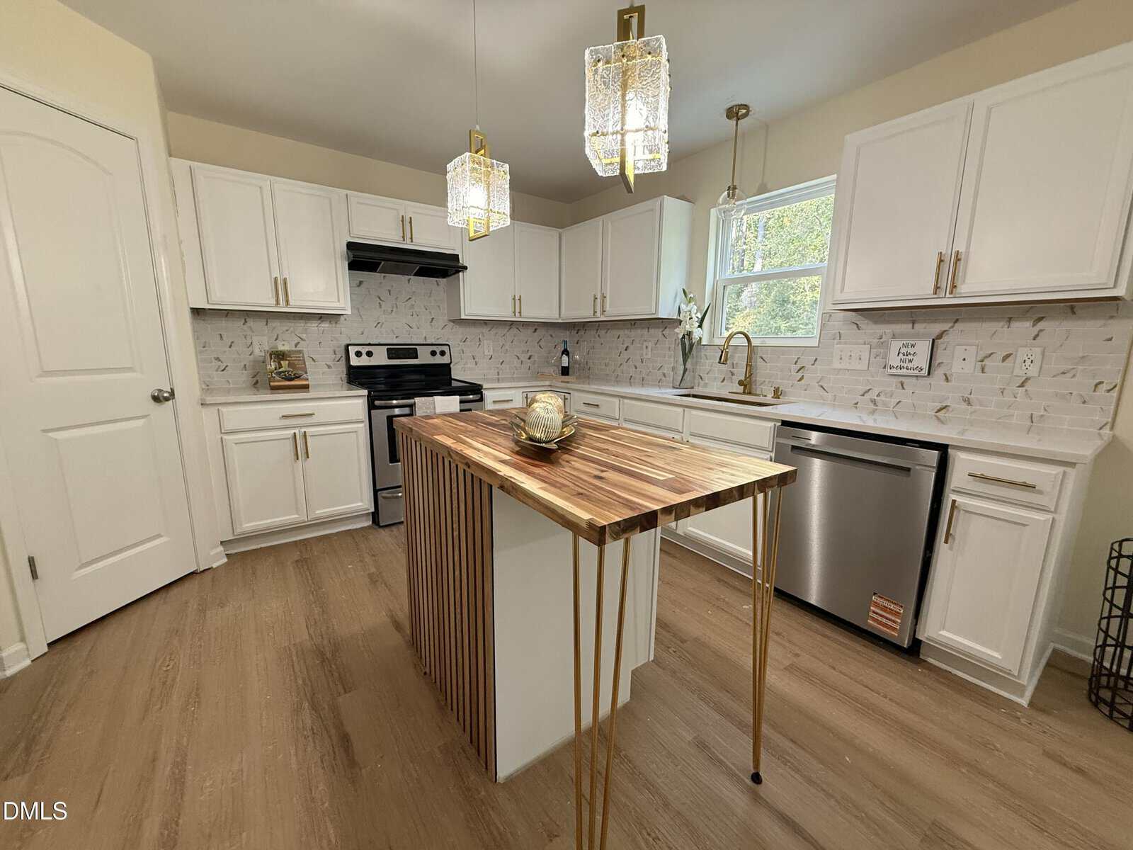 A modern kitchen with white cabinets, stainless steel appliances, a wood and gold island, pendant lights, a tile backsplash, and light-colored wood flooring. A window above the sink lets in natural light.