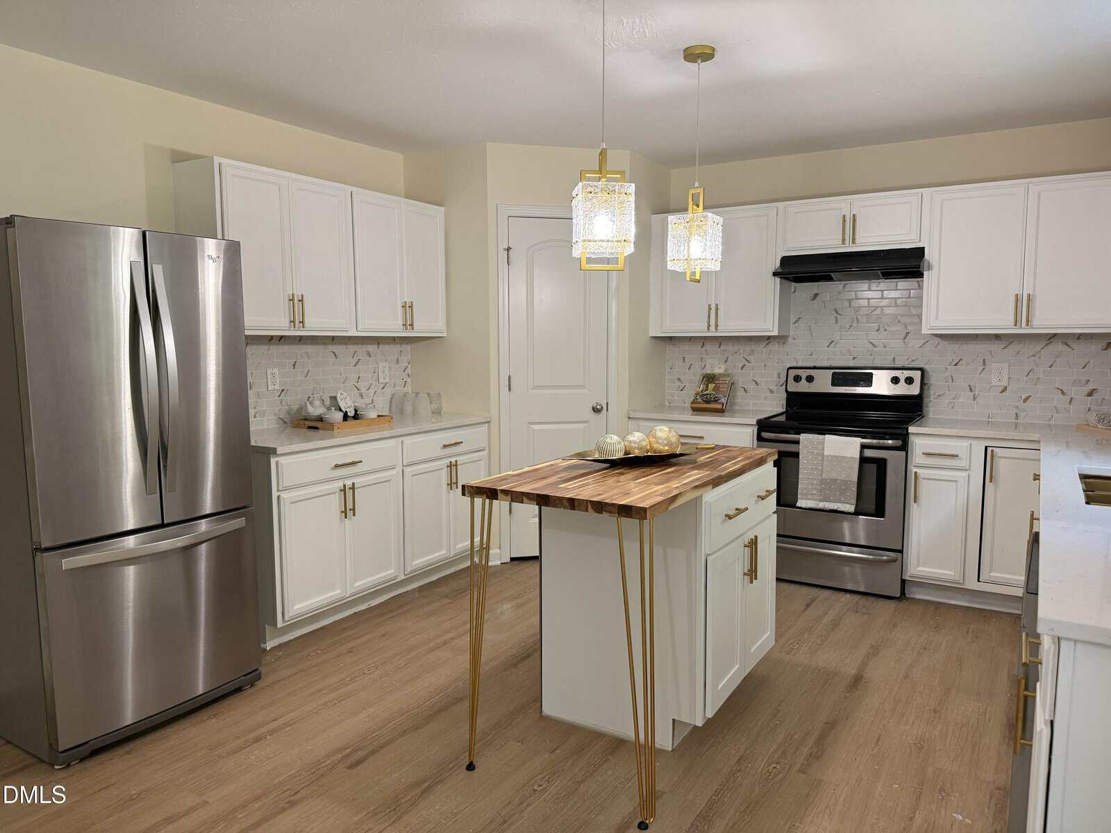 Modern kitchen with white cabinets, stainless steel appliances, a wooden island with gold legs, light wood flooring, and two pendant lights hanging above the island. Marble-patterned backsplash and countertop are visible.