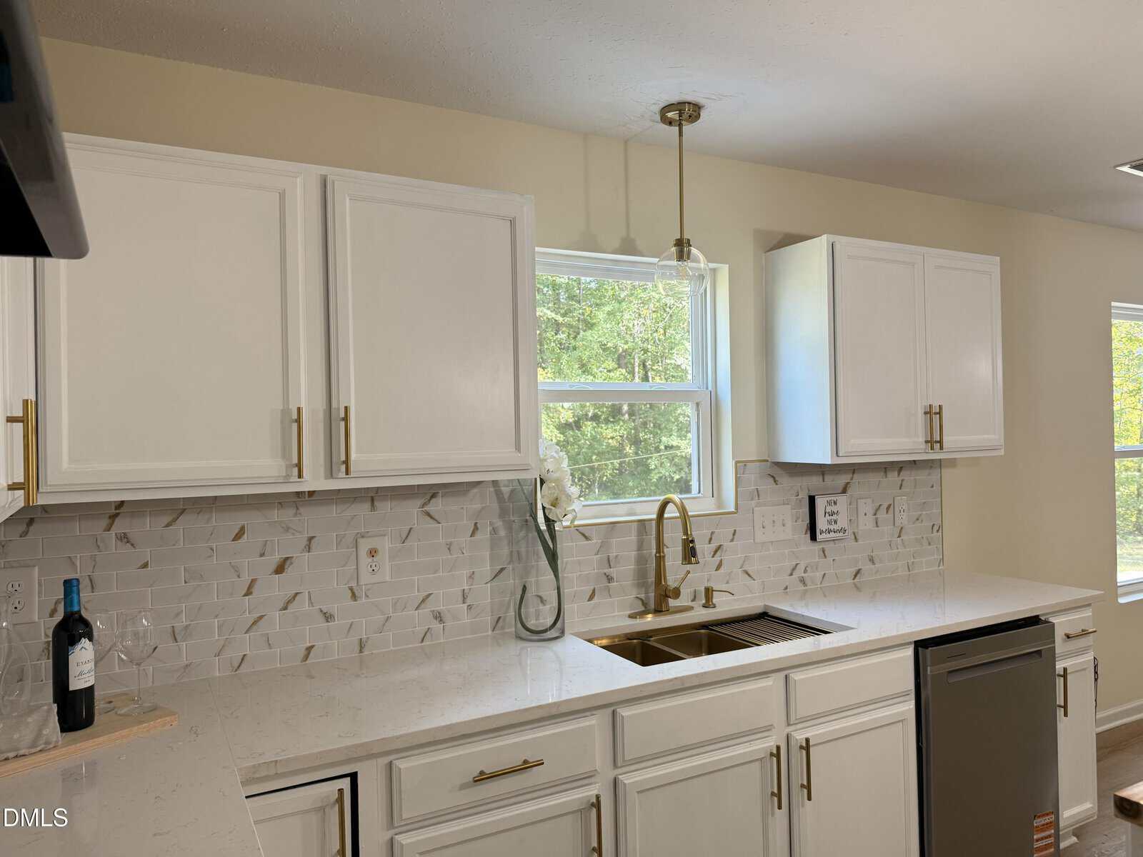 A modern kitchen with white cabinets, gold hardware, a white marble countertop, geometric tile backsplash, a stainless steel dishwasher, gold faucet, wine bottle, vase with white flowers, and a window above the sink.