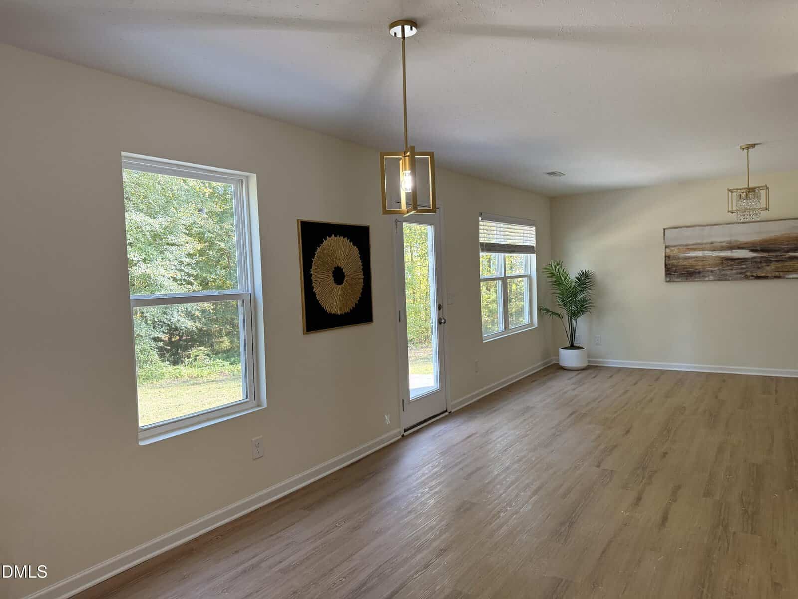 A bright room with light wood flooring, cream-colored walls, large windows, a glass door, two modern pendant lights, wall art, and a potted plant. Trees are visible outside through the windows.