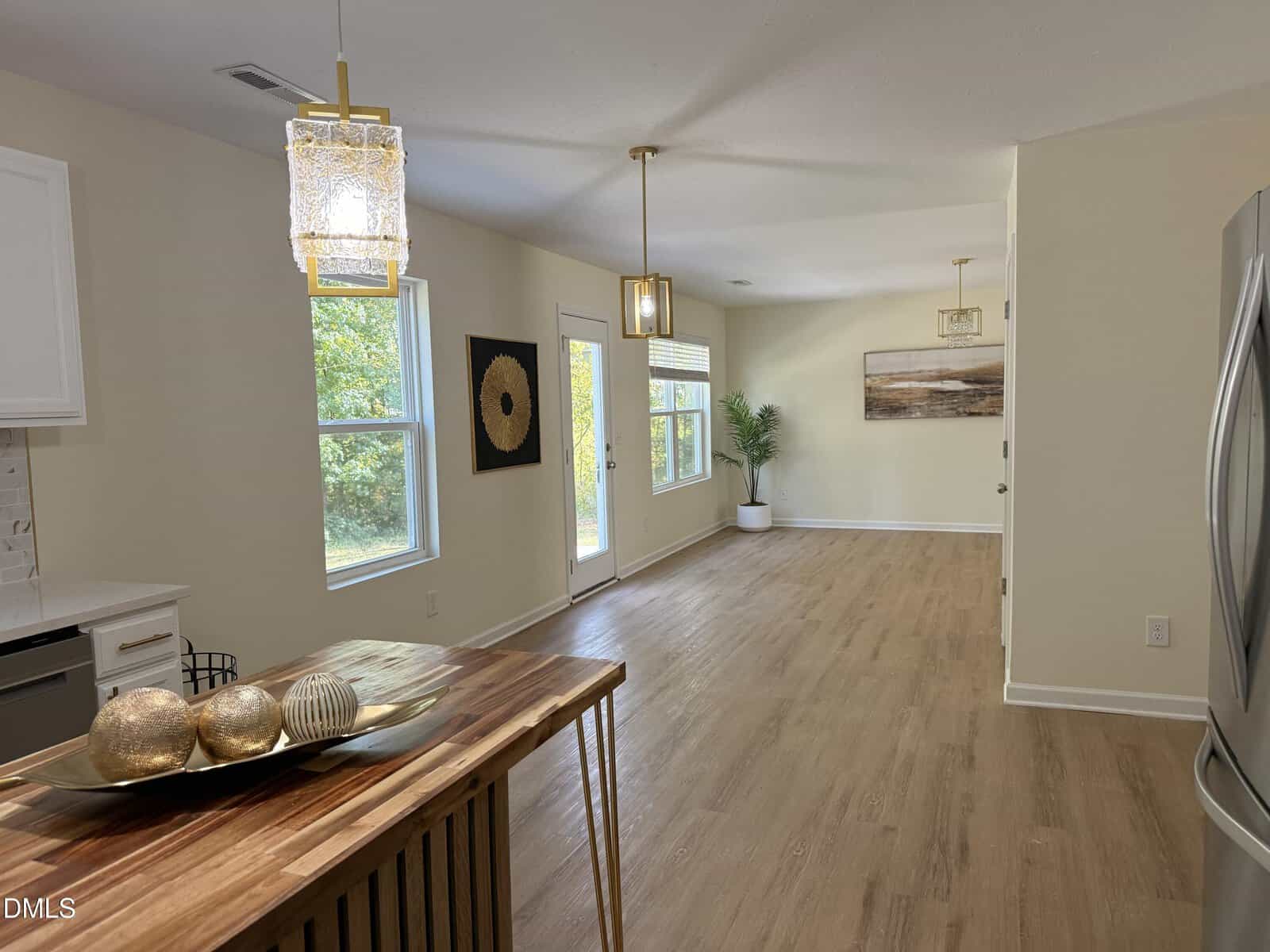 A bright, open-concept interior with light wood flooring, cream walls, pendant lights, large windows, a door to the outside, and minimal decor including a plant and a decorative bowl on a wooden island.