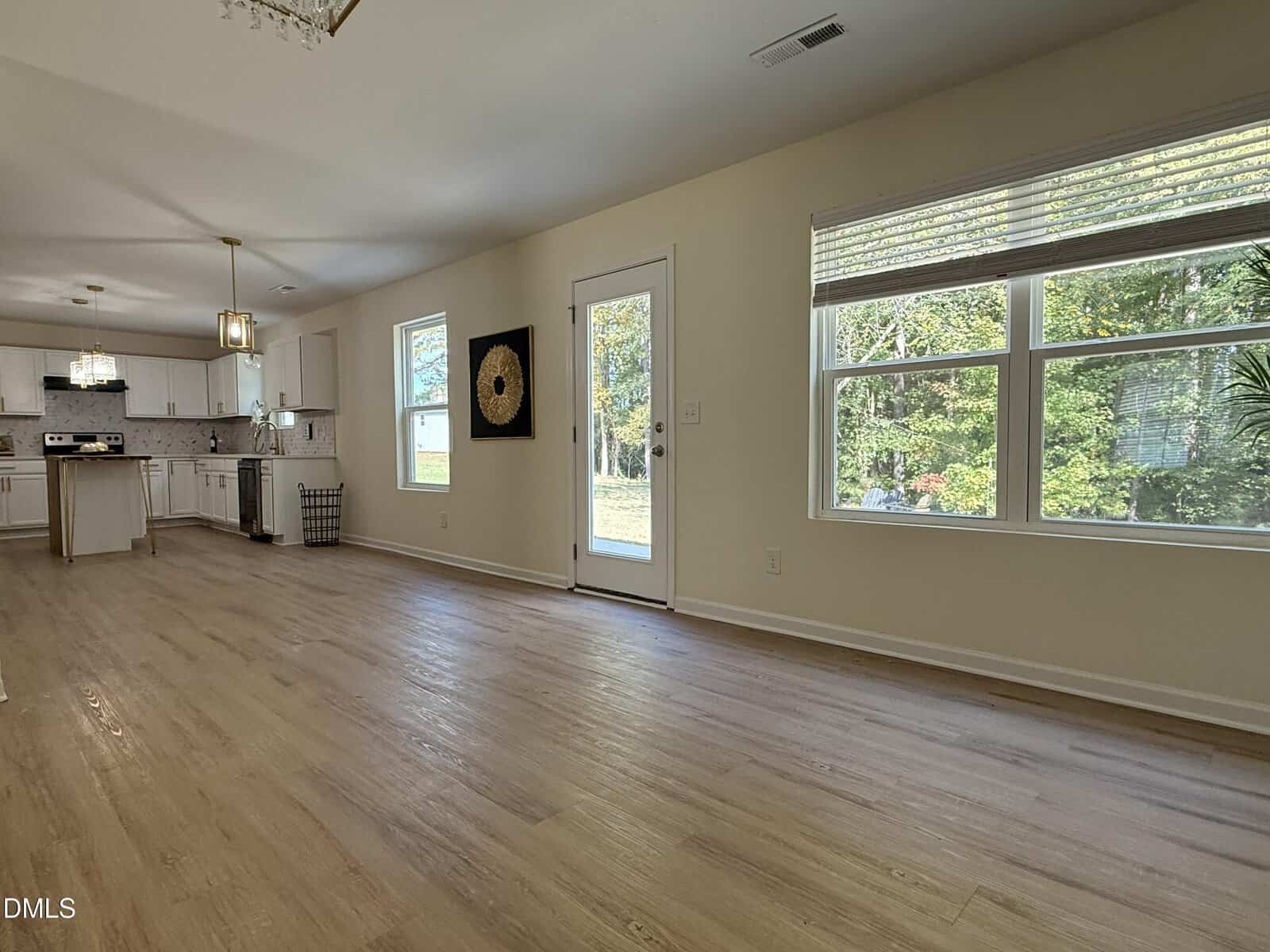 Spacious, empty living room with light wood flooring, white walls, large windows, glass door to backyard, and an open kitchen with white cabinets and stainless steel appliances in the background.