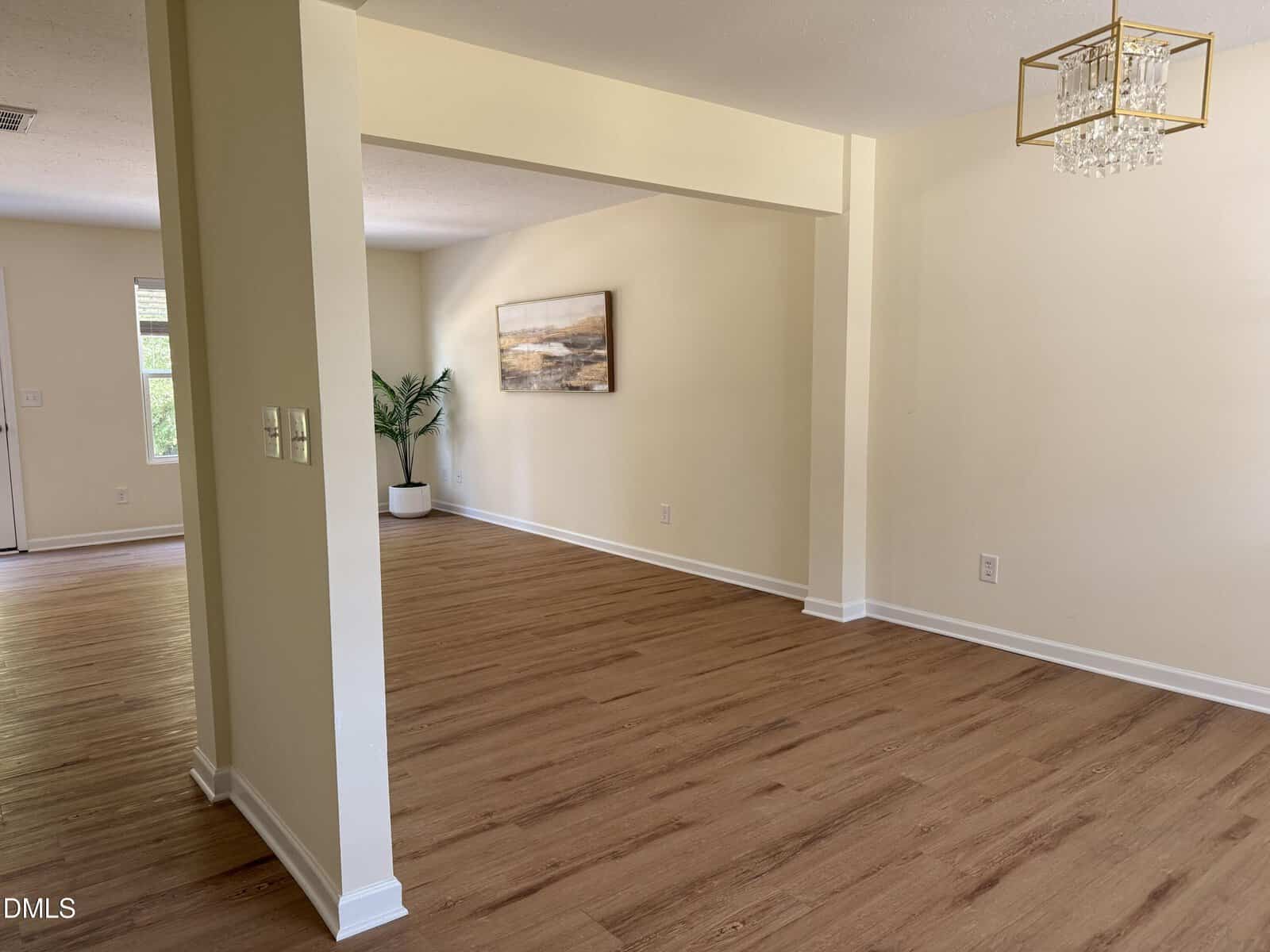 Spacious, unfurnished room with light wood flooring, cream walls, a modern gold chandelier, and a potted plant in the corner. A landscape painting hangs on the far wall. Natural light brightens the space.