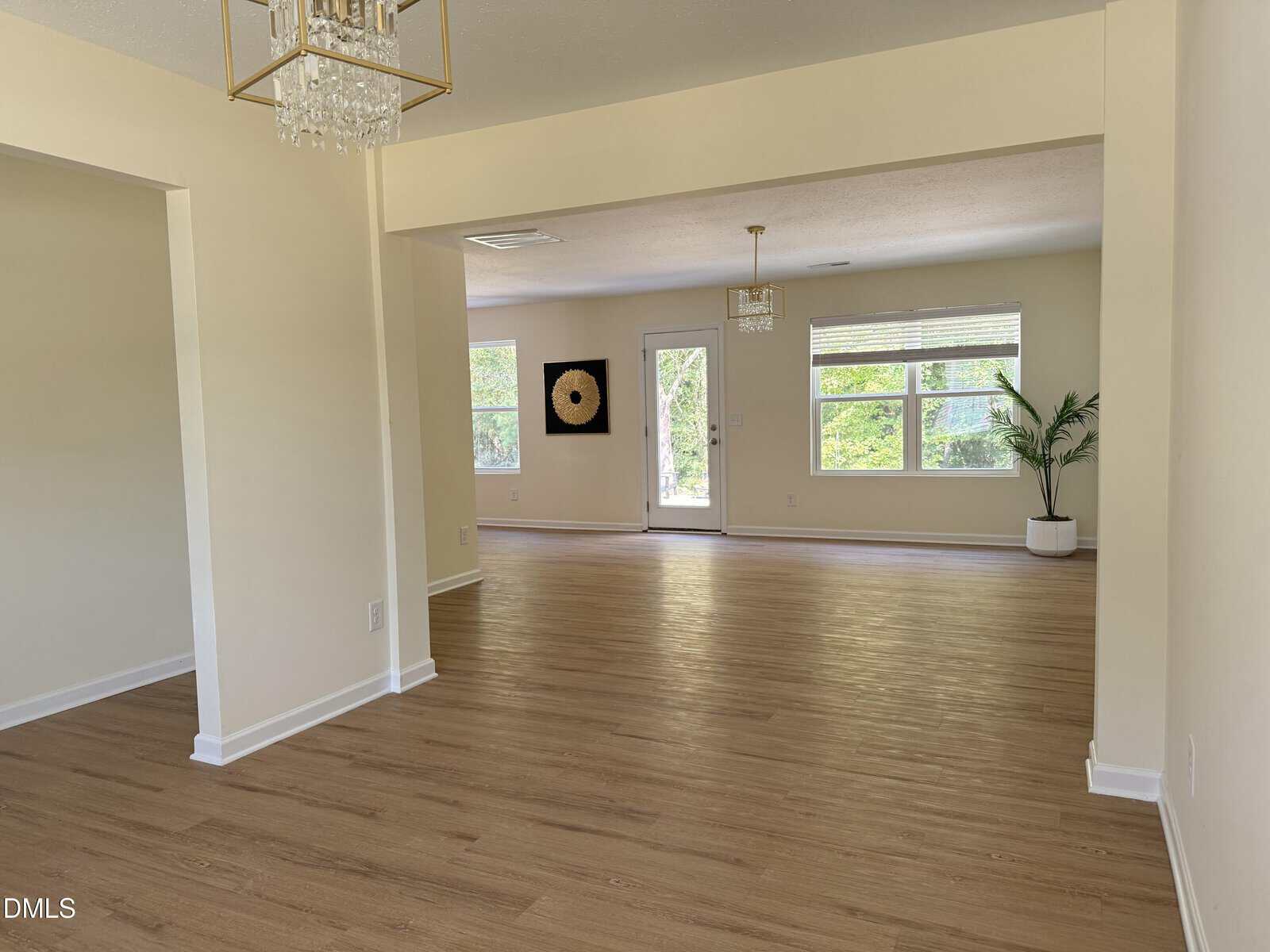 Spacious, empty living area with light wood floors, cream walls, large windows, a glass door, modern chandeliers, and a potted plant by the window. Natural light fills the room.
