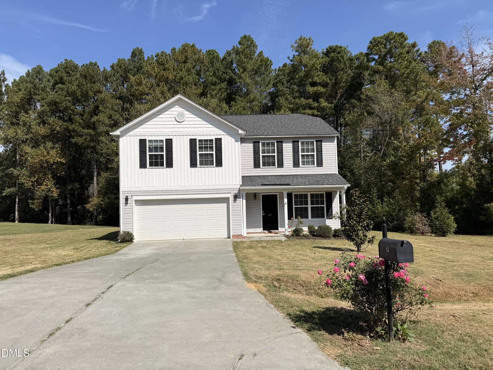 A two-story white house with black shutters and a gray roof, a double garage, a concrete driveway, a front lawn with some bushes, and a mailbox with pink flowers, surrounded by tall trees under a blue sky.