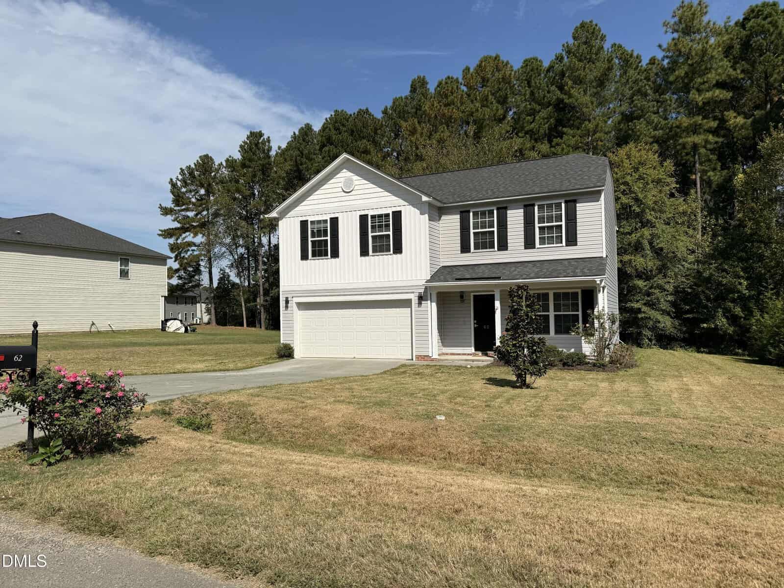 Two-story white house with black shutters and a gray roof, attached two-car garage, front porch, and a well-kept lawn with some bushes and trees, set against a backdrop of tall pine trees under a blue sky.