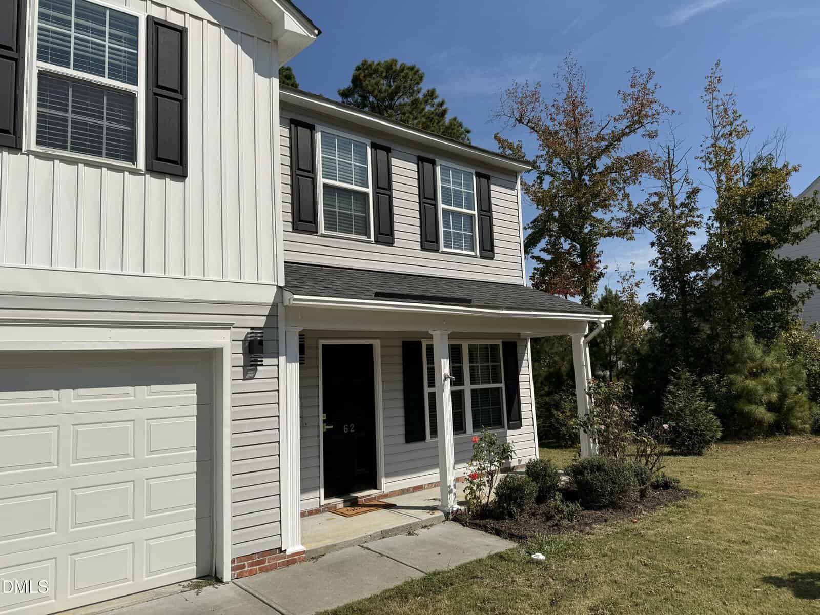 Two-story white house with black shutters and a single-car garage. The front porch has white columns, a black door, and some landscaping with bushes and a small tree. Blue sky and trees are in the background.