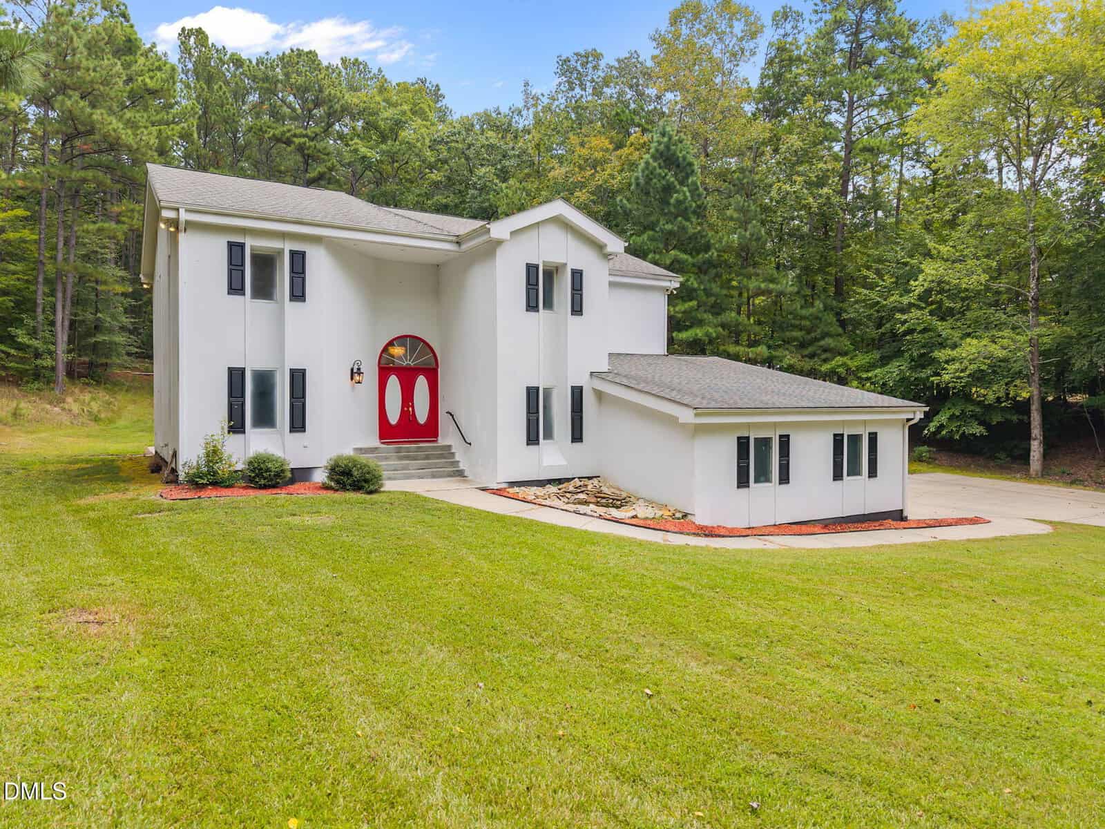 A white two-story house with a red double front door, attached single-story section, and concrete driveway, surrounded by grass and trees under a blue sky.