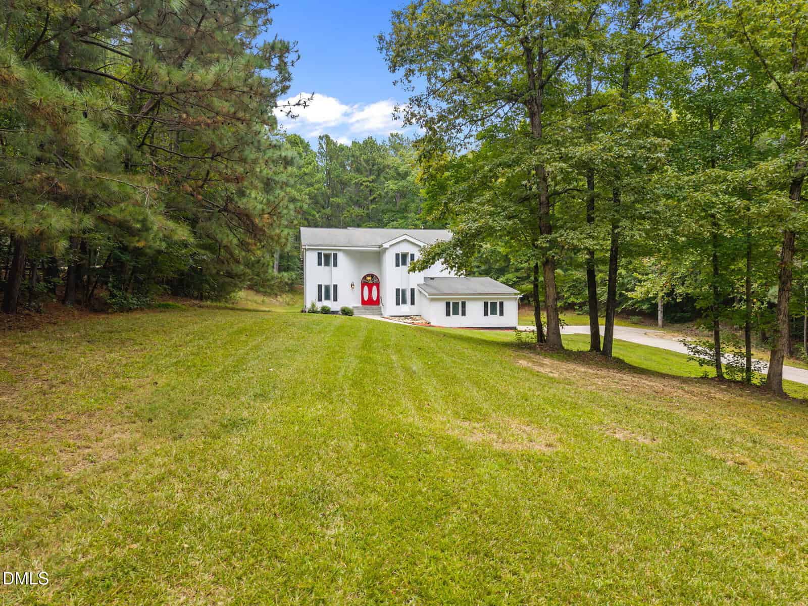 A two-story white house with a red front door and an attached garage is surrounded by trees and sits on a large, sloped grassy lawn under a partly cloudy sky.