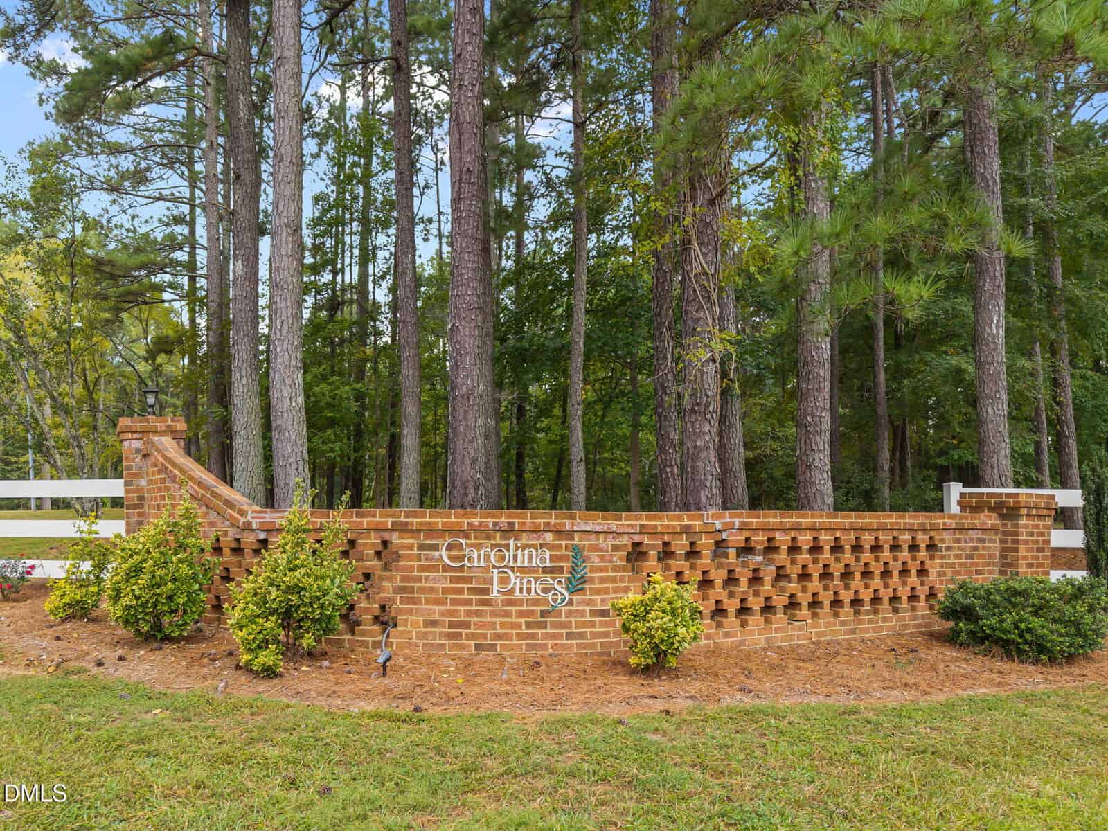 A brick sign with "Carolina Pines" written on it stands in front of a pine forest, bordered by shrubs and pine straw mulch, with a white fence partially visible on the left.