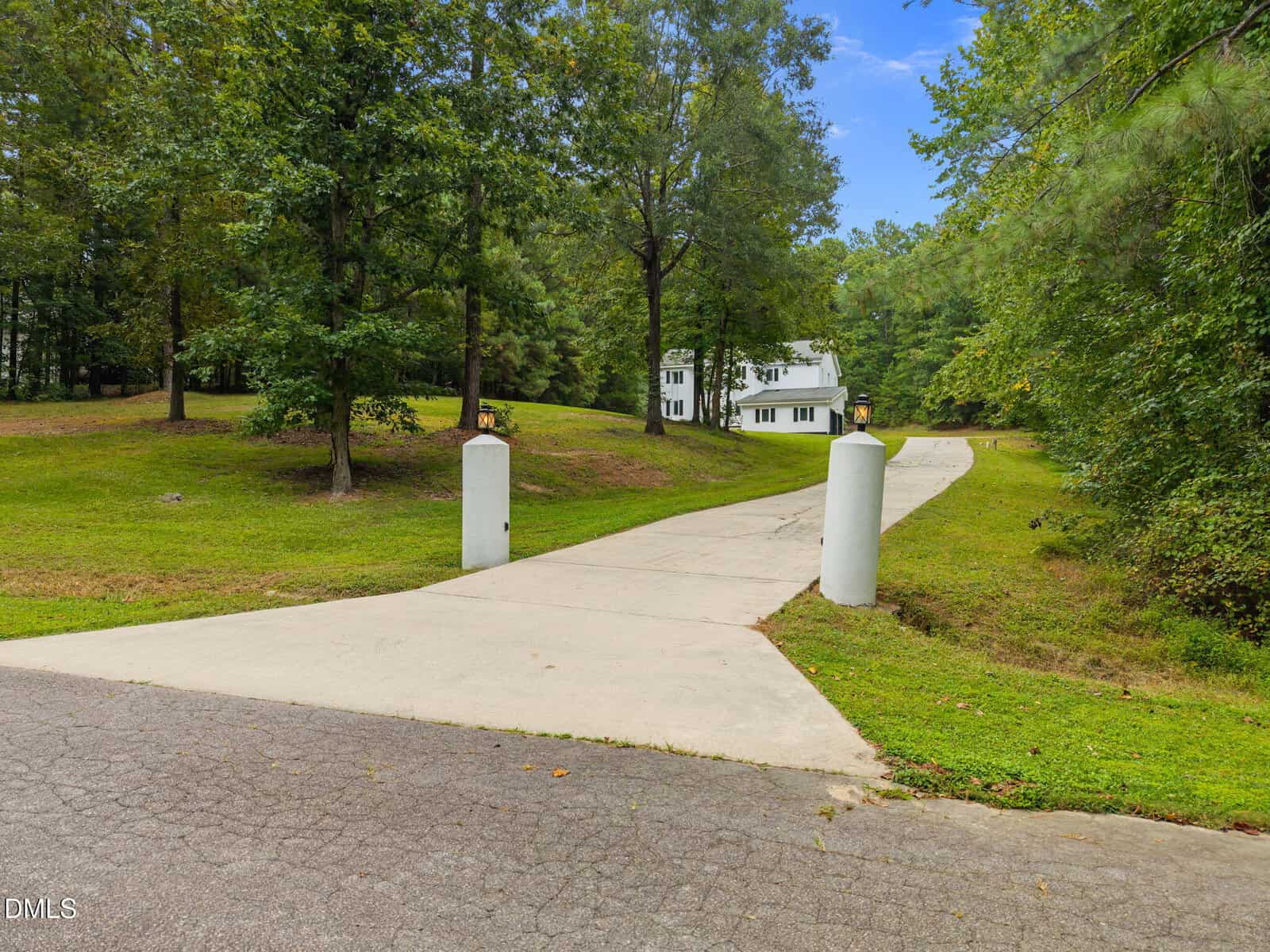 A long concrete driveway with two white pillars at the entrance leads through a grassy, wooded area to a white two-story house in the background. The sky is clear and blue.