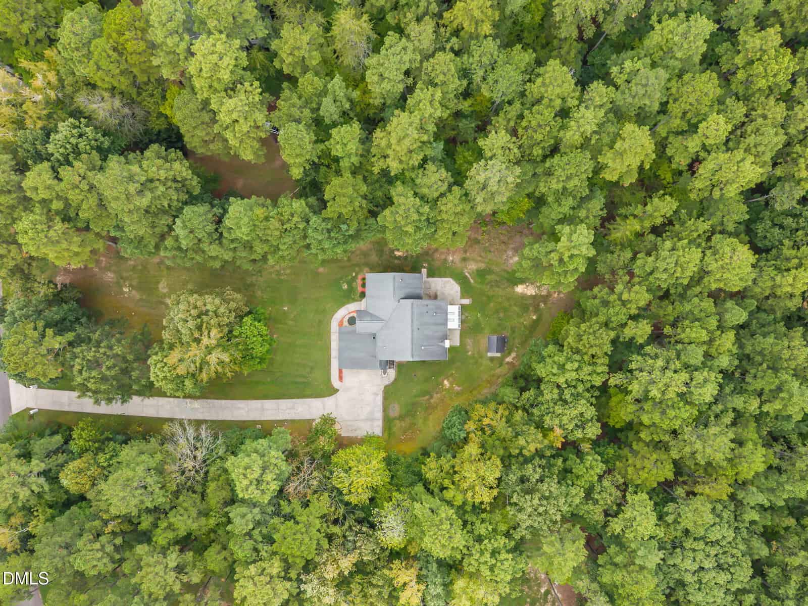Aerial view of a gray house with a curved driveway surrounded by dense green trees, set on a large grassy lot with a small detached structure nearby.