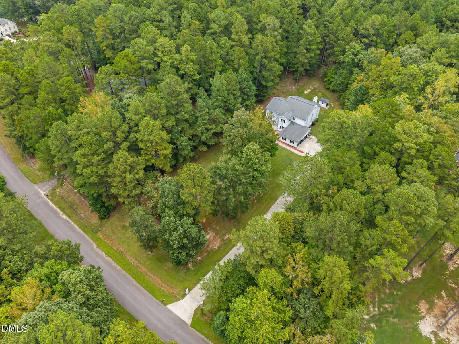 Aerial view of a two-story house with a white exterior and gray roof, surrounded by dense green trees, located near a curved paved road in a forested residential area.