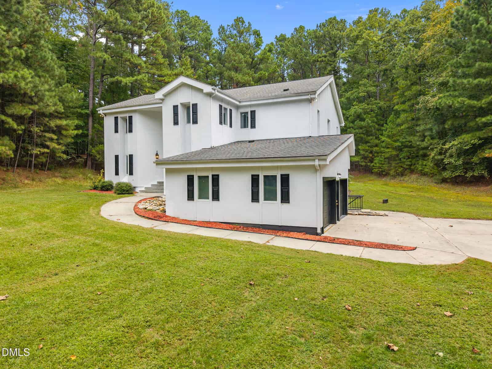 A modern two-story white house with black window shutters sits on a spacious, well-maintained lawn. The property is surrounded by tall trees and has a paved driveway curving around the home.
