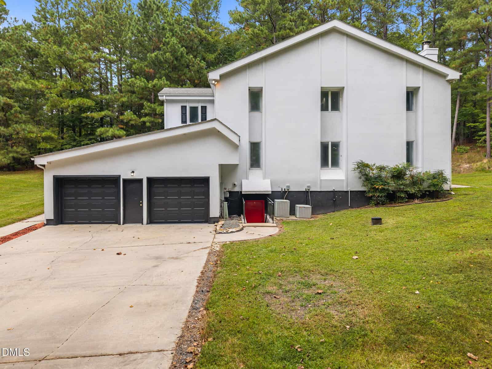 A modern white house with two tall vertical windows, a double garage, a driveway, and surrounding green lawn. Trees are in the background and HVAC units are visible against the building's side.