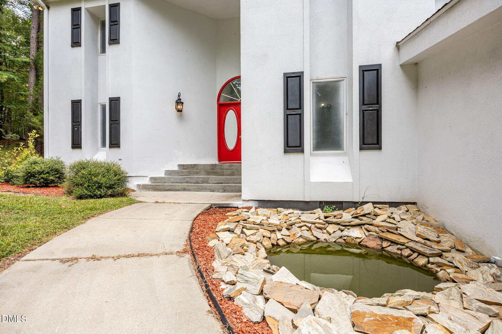 A curved sidewalk leads to a white house with a bold red front door, black shutters, and a small pond with rocks beside the entrance. Shrubs and mulch border the walkway.