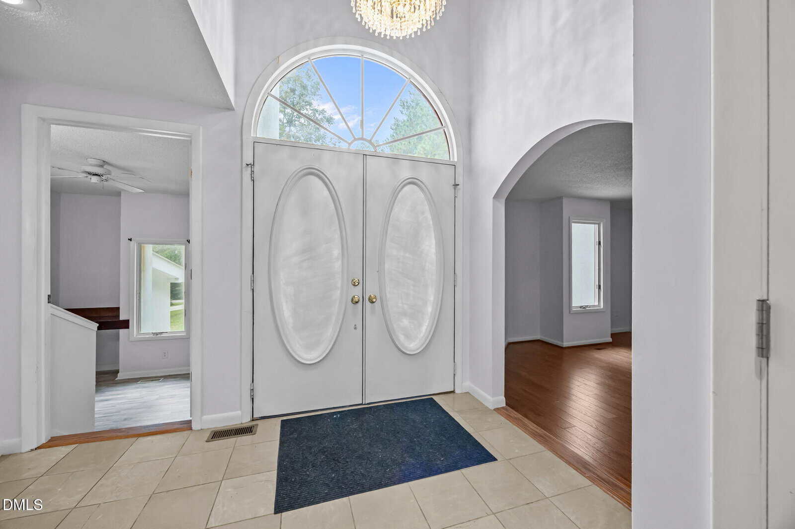 Bright foyer with double oval-front doors, chandelier, tile floor, and large arched window. Adjacent rooms include a wood-floored living area and a carpeted room visible through arches.