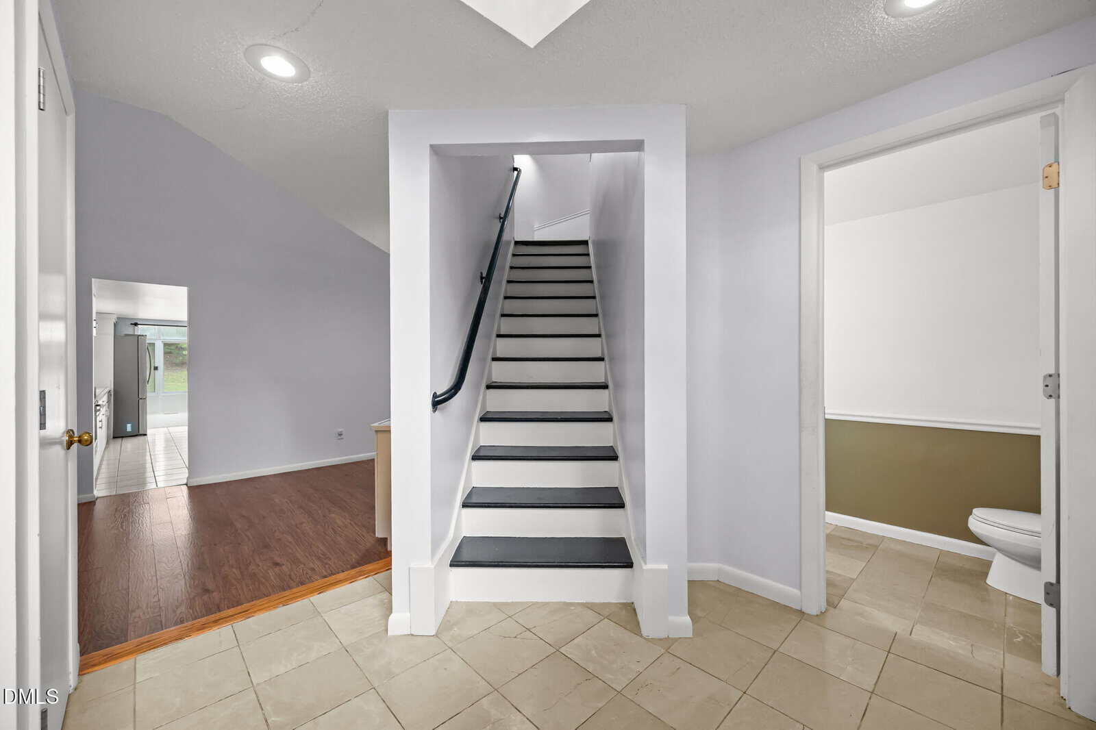 A modern entryway with beige tile flooring, a white staircase with dark handrails leading up, an open doorway to a room with wood floors, and a bathroom with a toilet visible on the right.