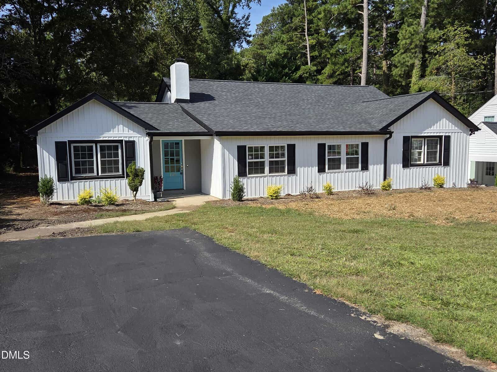 A single-story white house with black trim at 115 Red Bud Drive, Henderson features a turquoise front door, black roof, paved driveway, and several windows. Small shrubs line the front with a lawn and trees in the background.