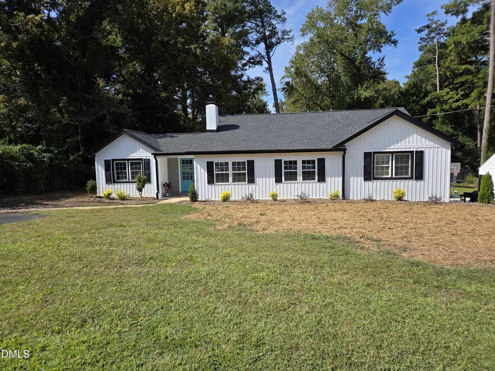 A single-story white house with black shutters and a teal front door sits on a spacious grassy lawn, surrounded by trees and small shrubs planted along the front.
