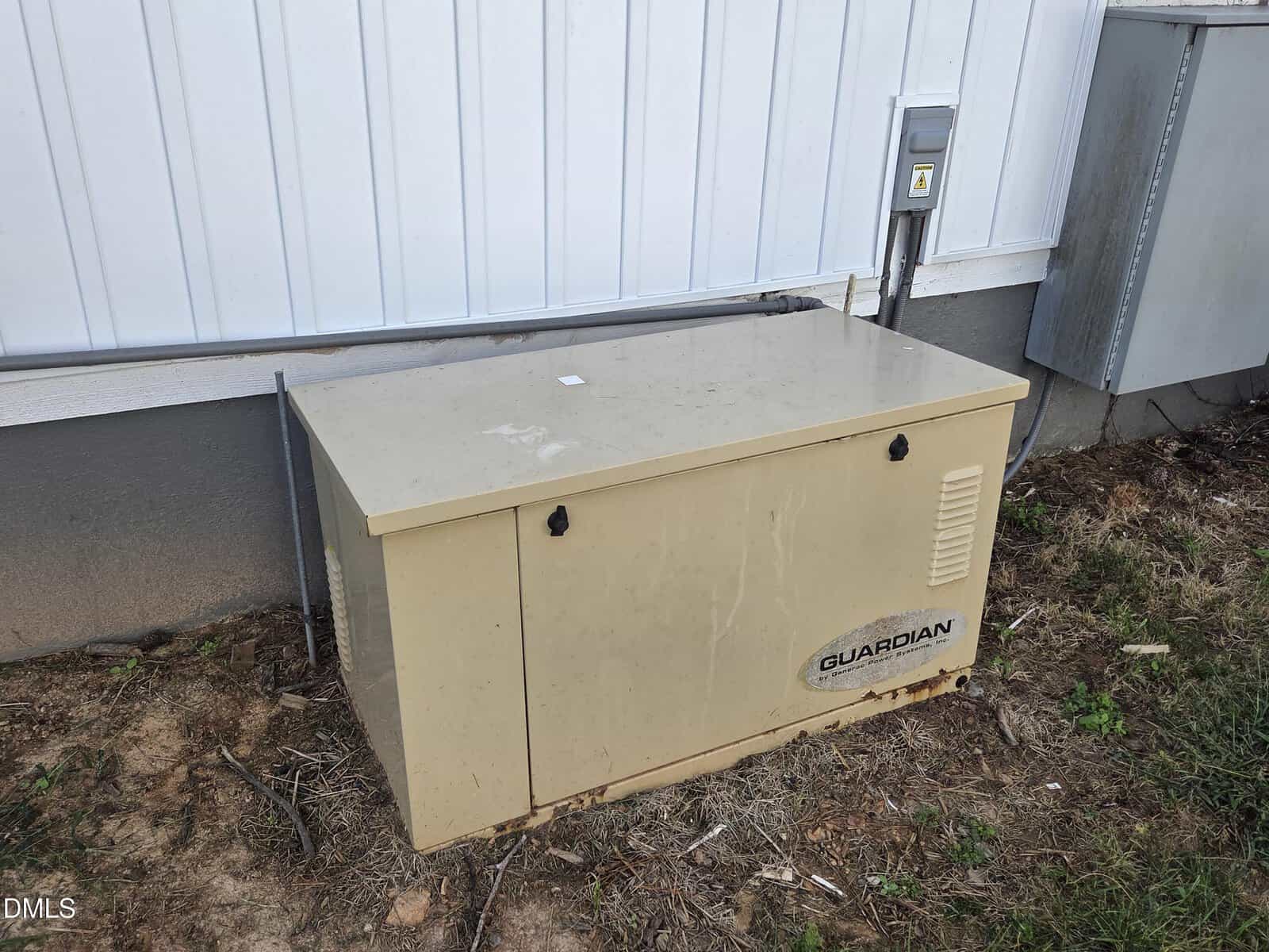 A beige Guardian generator sits on the ground outside a building with a white metal wall and gray foundation. Electrical panels and conduit are attached to the wall nearby.