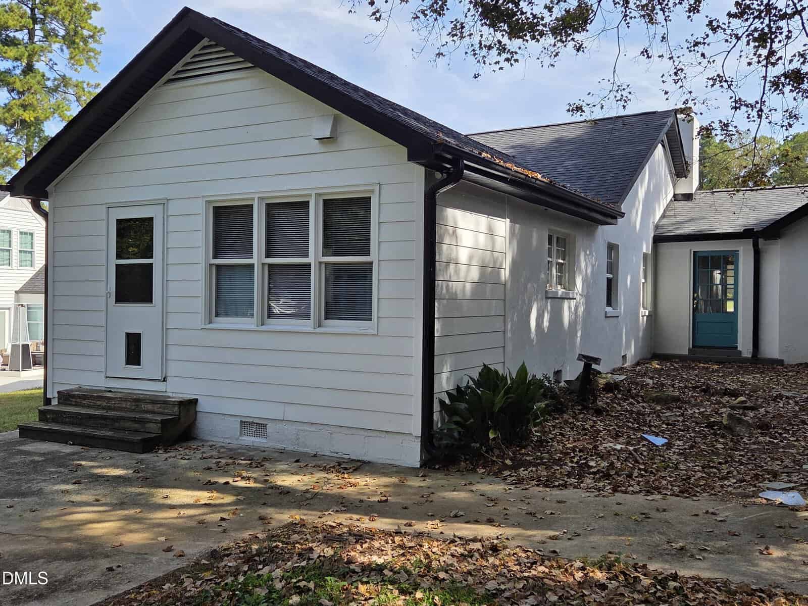 A white, single-story house with black trim, a small front porch with steps, and several windows. Fallen leaves cover the ground and driveway. There is a side door visible, and trees in the background.
