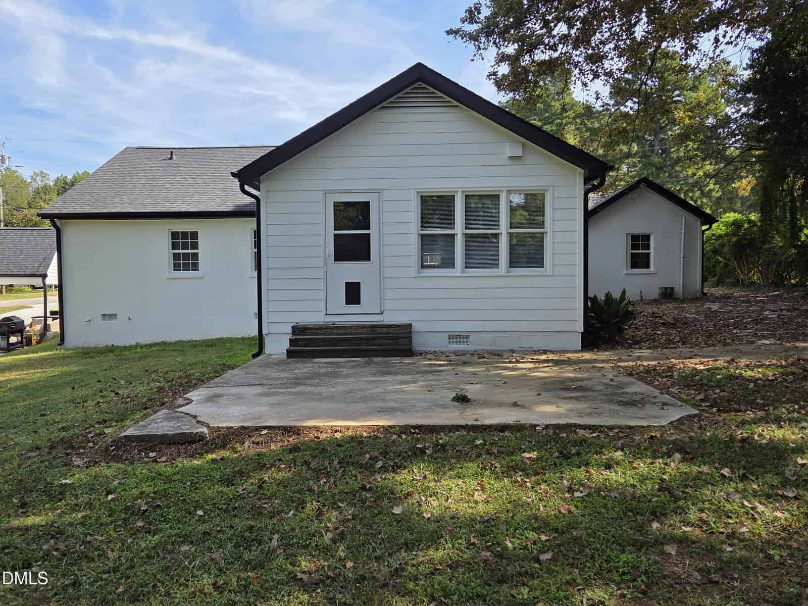 A small white house with black trim sits on a grassy yard. There are three windows and a door on the back side, concrete steps, and a concrete patio. Trees and another building are visible in the background.
