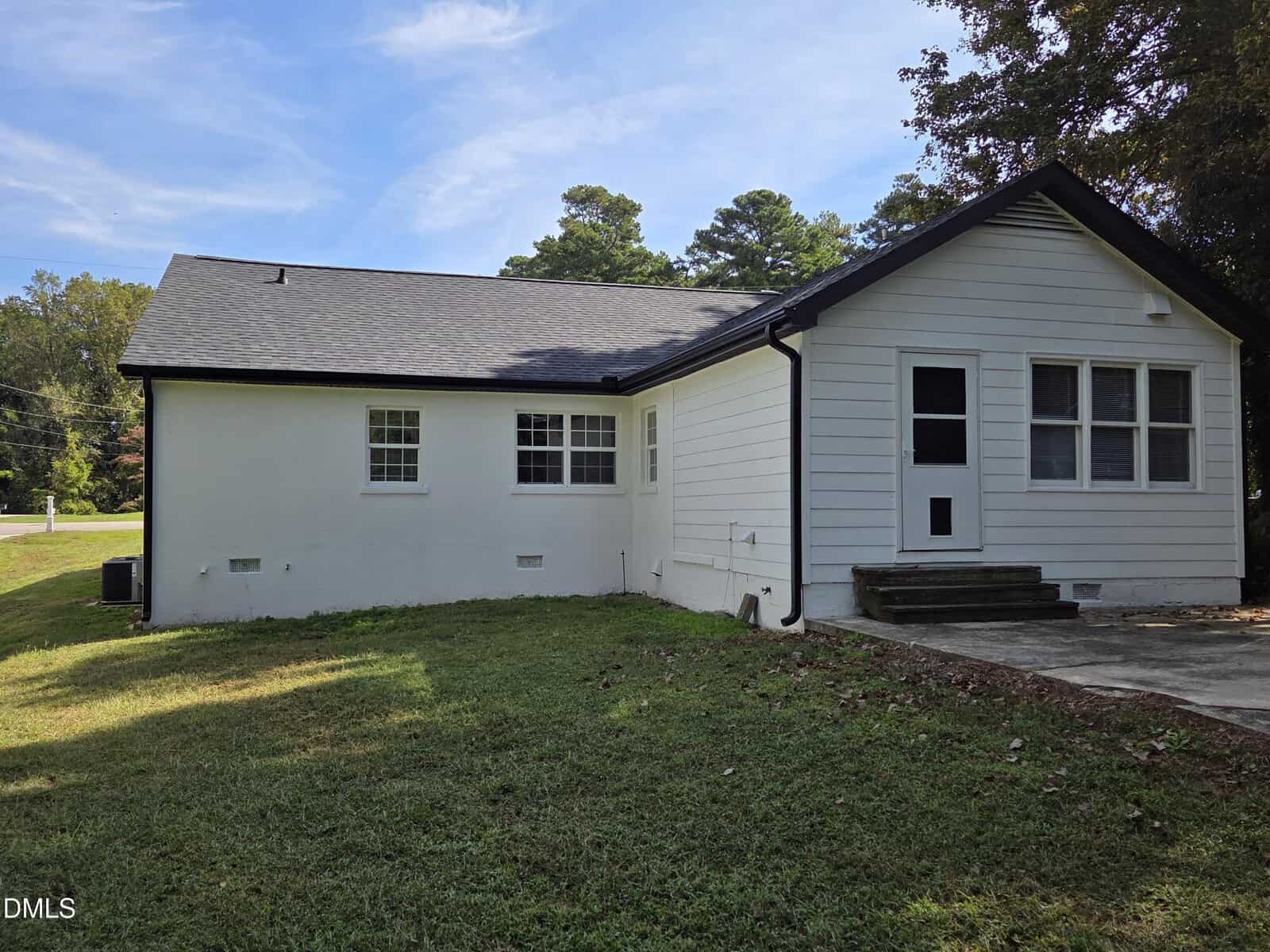 A white, single-story house with black trim, multiple windows, a dark shingled roof, concrete steps, and a grassy yard under a partly cloudy sky. Trees and a paved area are in the background.