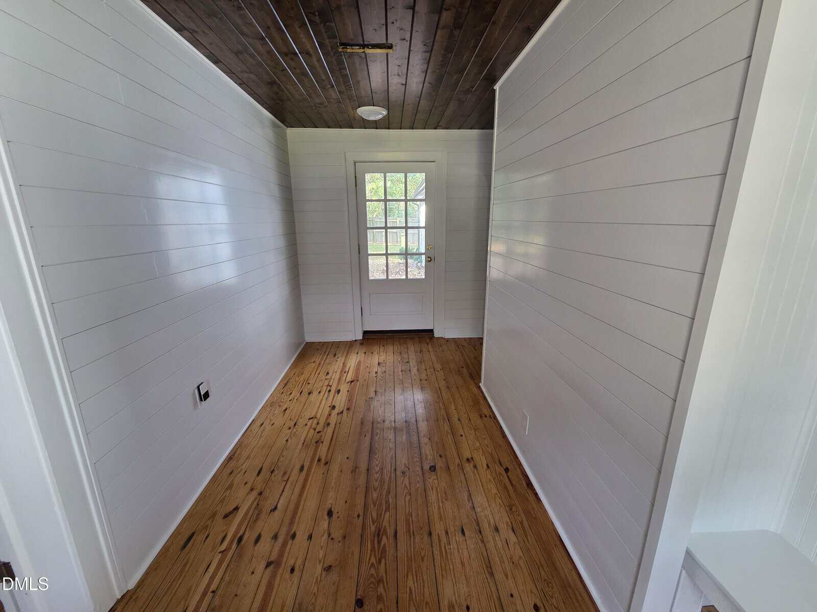 A narrow room with white shiplap walls, wood floor, and a dark wood ceiling. A glass door at the end lets in natural light. The room appears empty and freshly renovated.